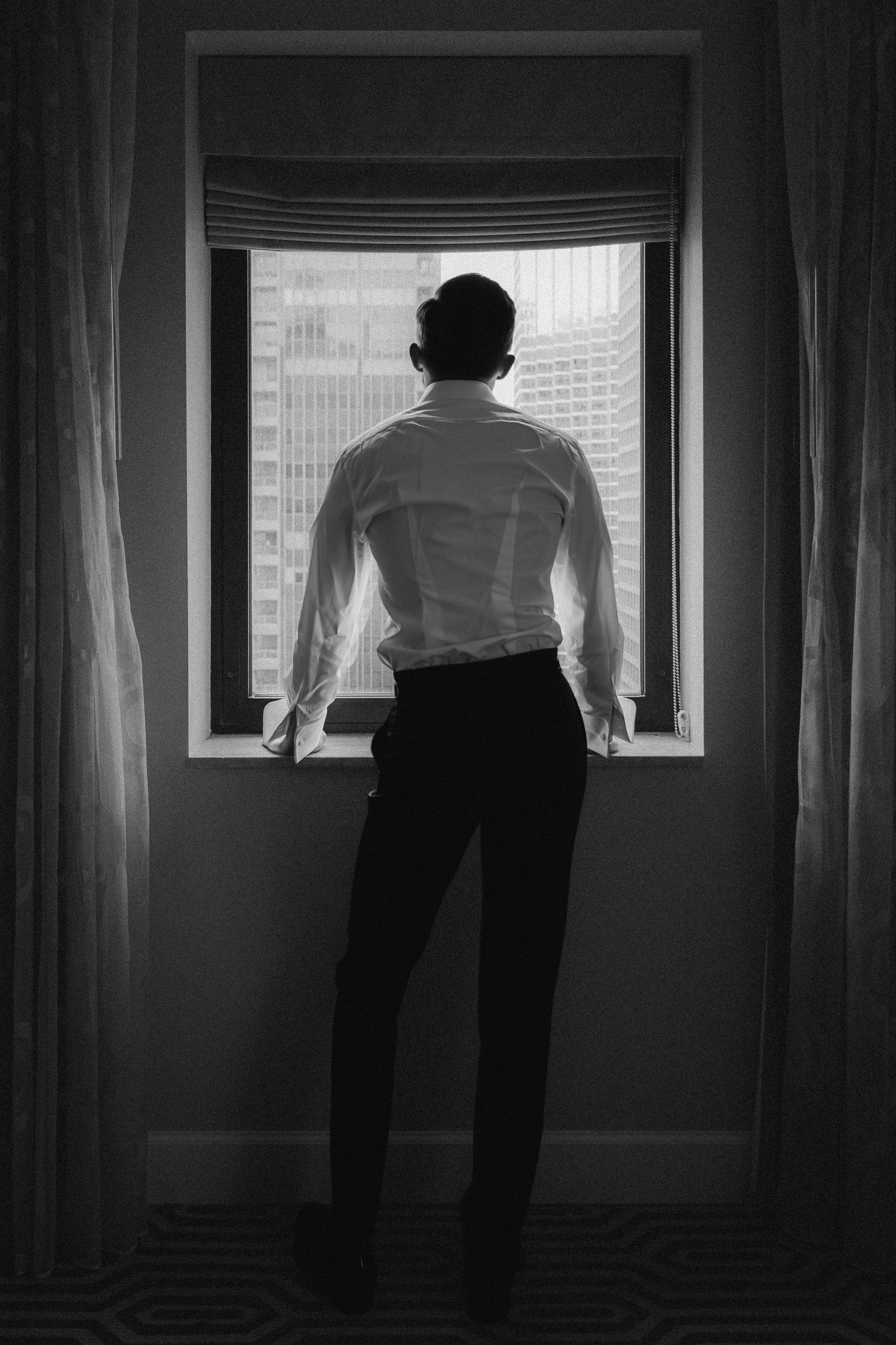 Man in white dress shirt and black pants stands gazing out hotel window at city skyline in black and white photo.