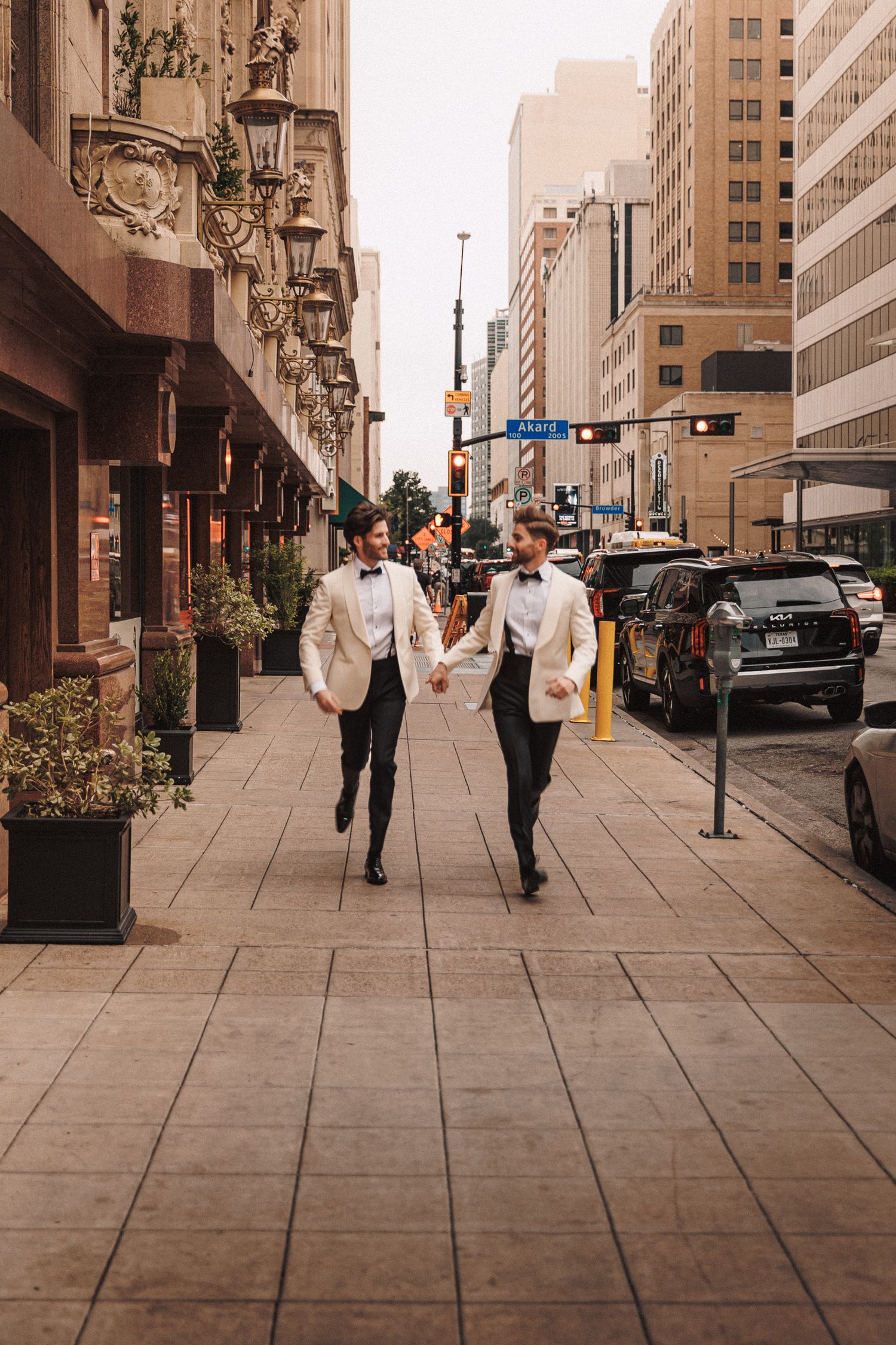 Two grooms in white tuxedo jackets hold hands walking down a city sidewalk on their wedding day.