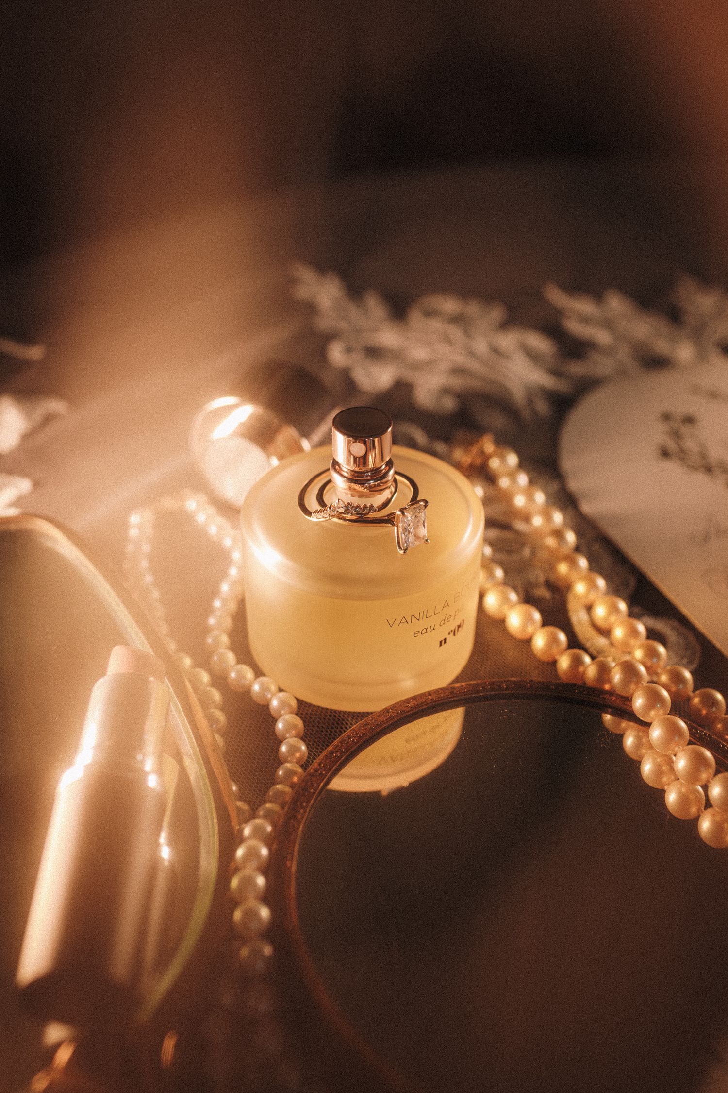 Golden perfume bottle with pearl necklace on mirrored vanity tray in warm romantic lighting.