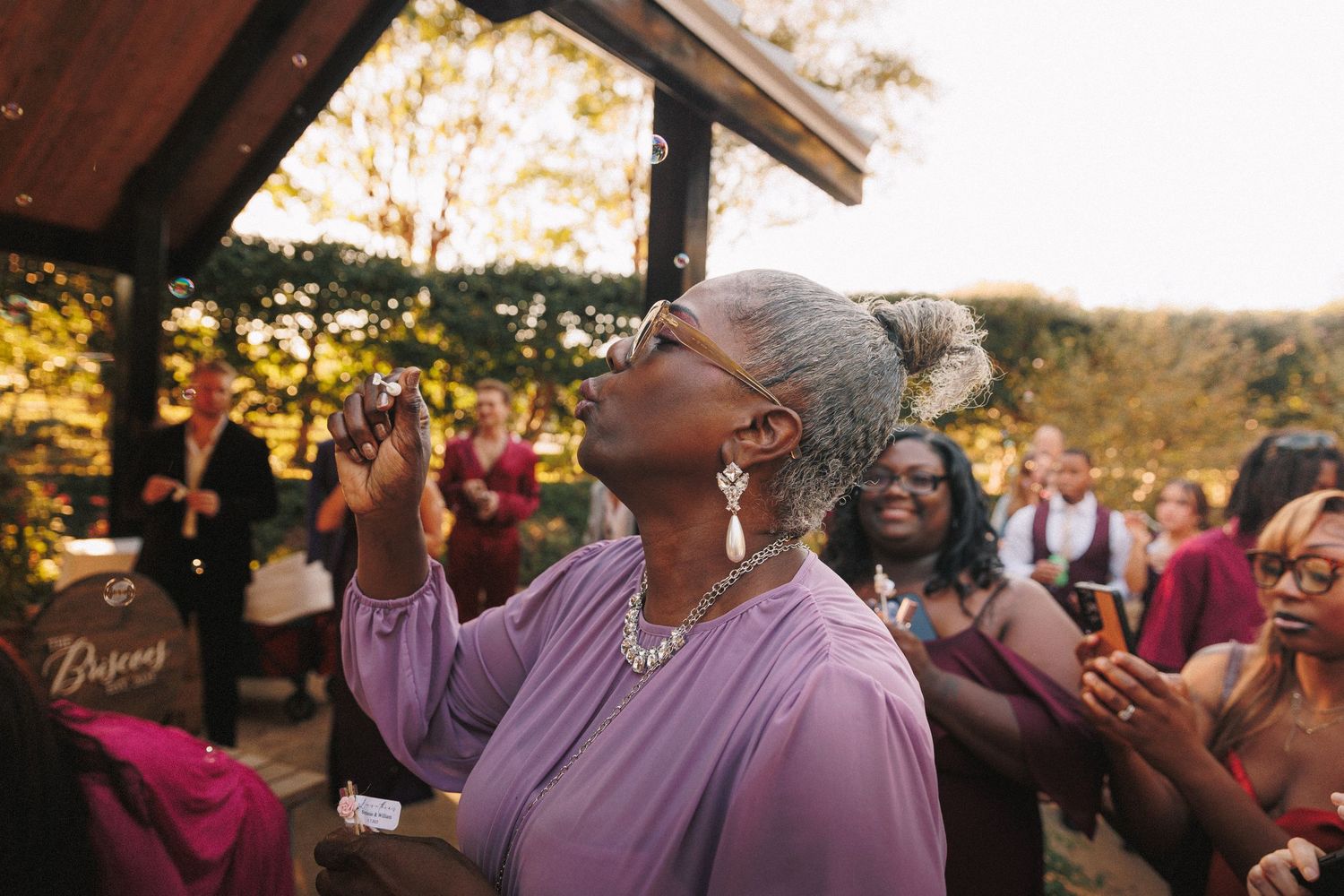 Elegant woman in lavender dress looking up joyfully at outdoor wedding celebration with guests in burgundy attire.