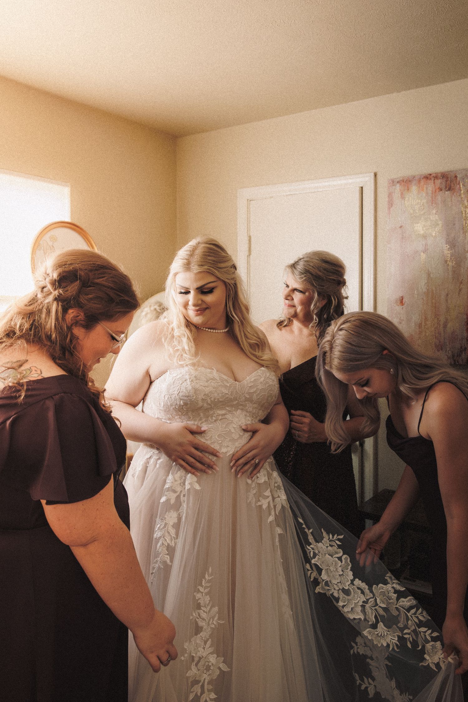 Bridesmaids in dark dresses helping bride into lace wedding gown in warm sunlit room.