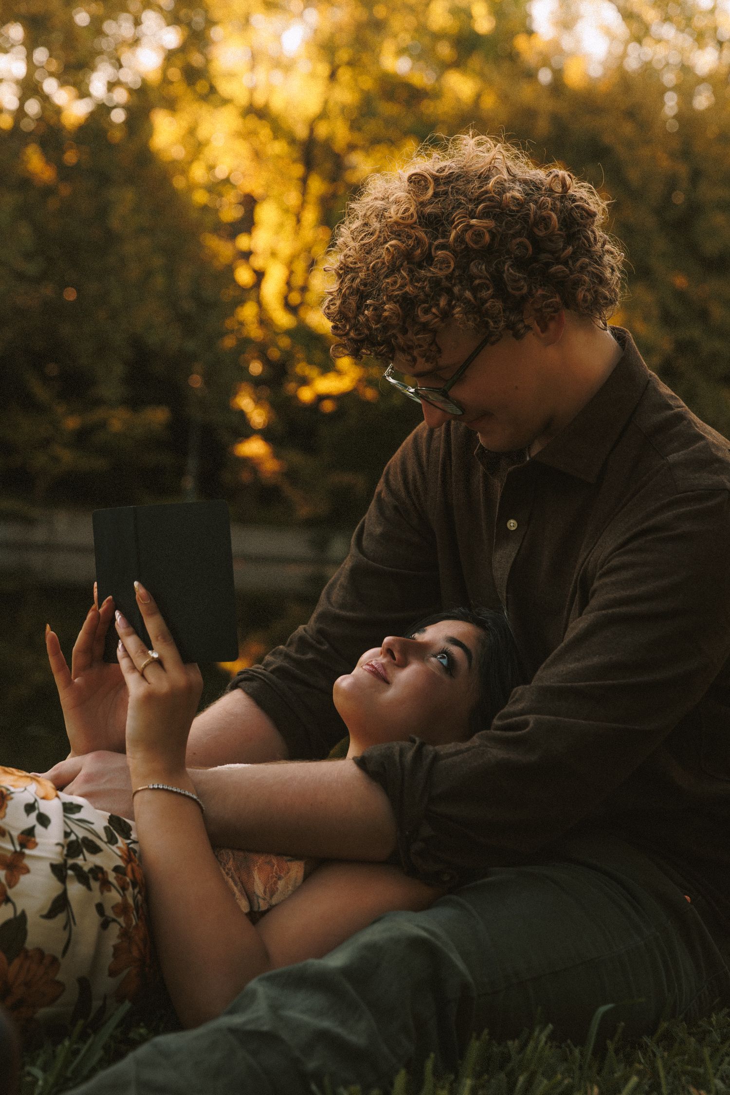 A couple relaxes together on grass during sunset with golden bokeh lighting in the background.