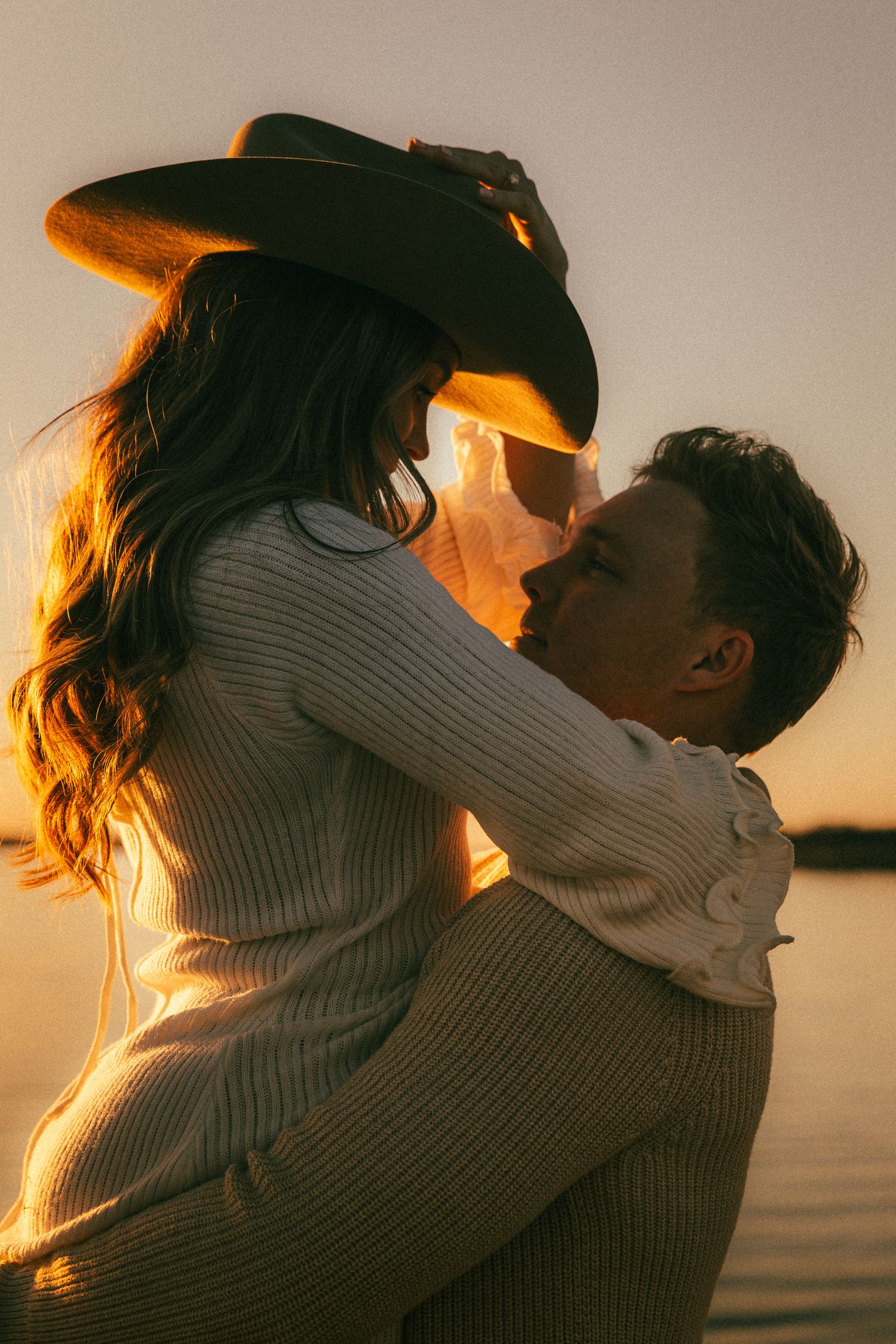 Silhouetted couple embracing at sunset by the water with golden light illuminating their tender moment.