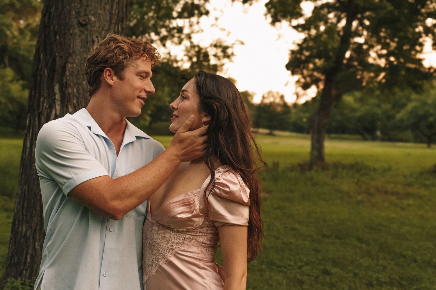 Couple embracing tenderly by a large tree trunk in a park during golden hour lighting.