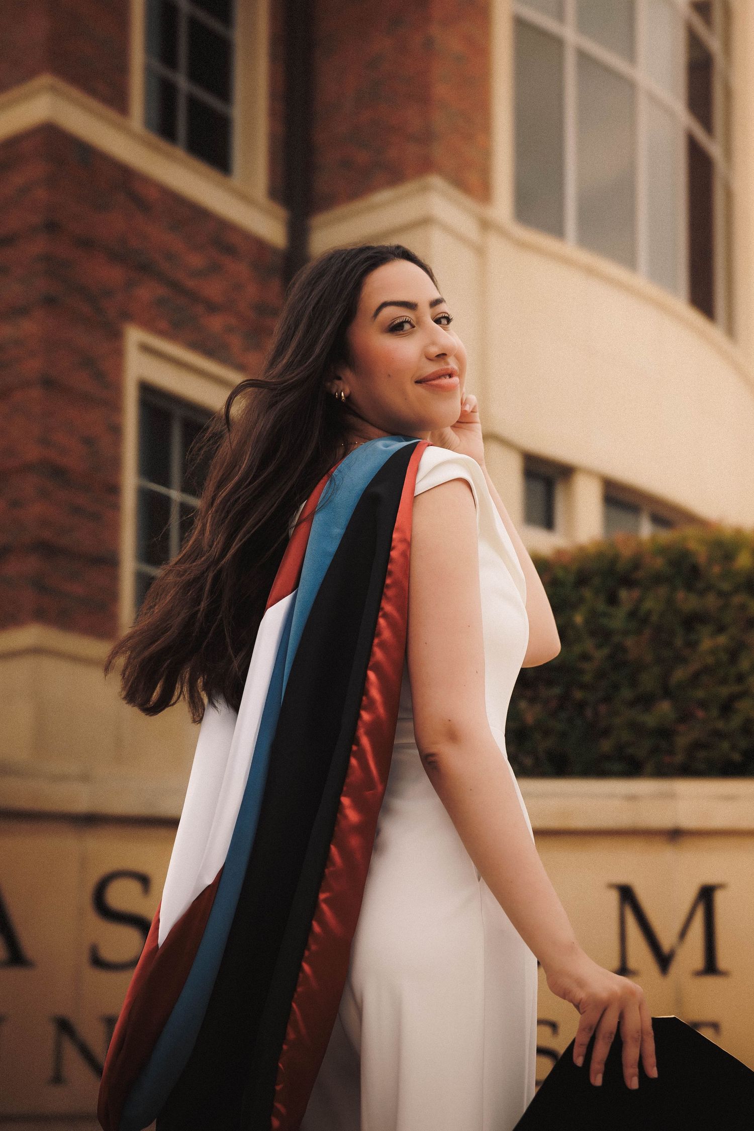 A graduating student in a white dress spins around joyfully outside a brick building with their hair flowing in the wind.