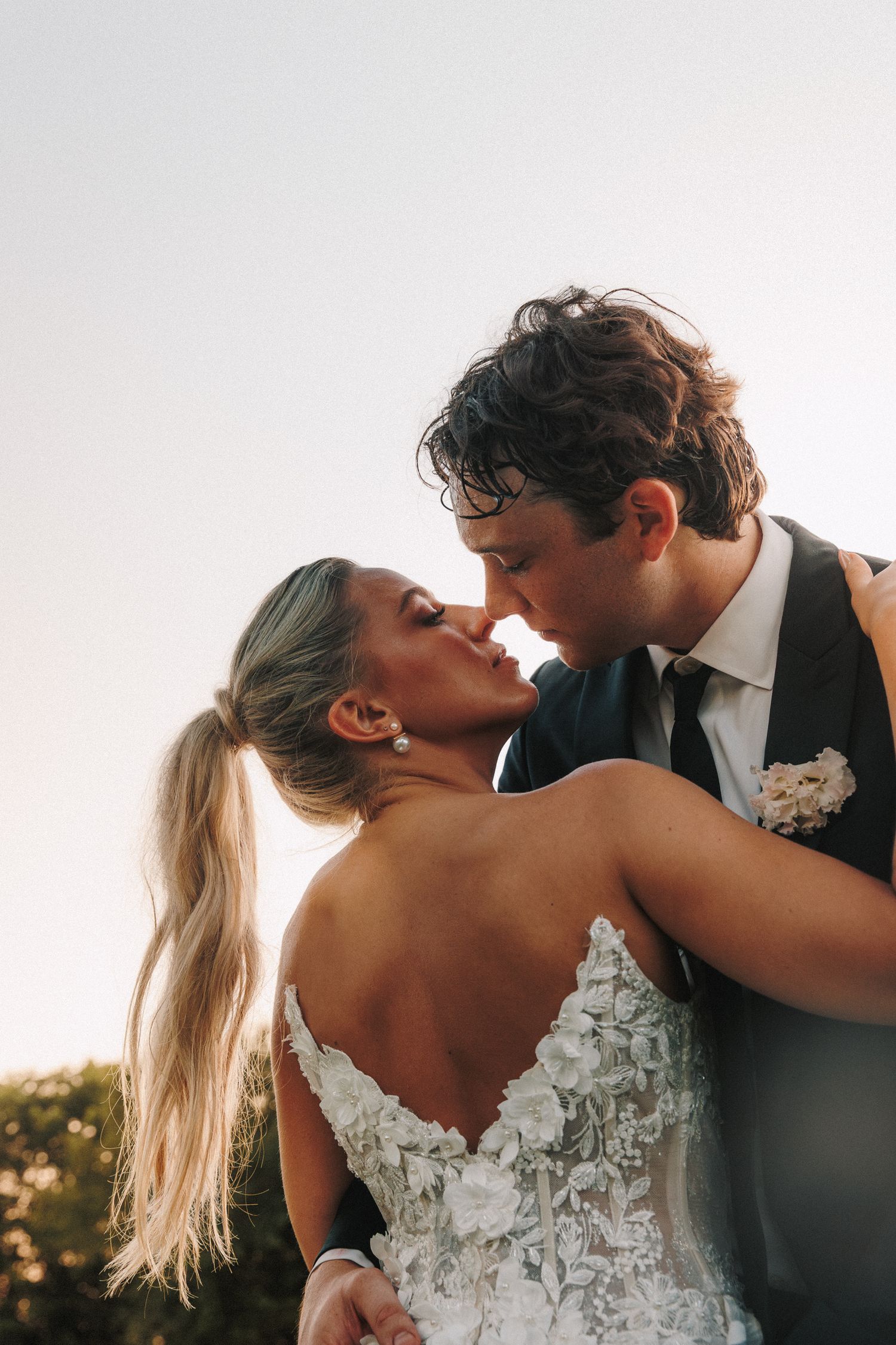 A romantic outdoor wedding scene with a loving couple in elegant attire kissing during sunset.