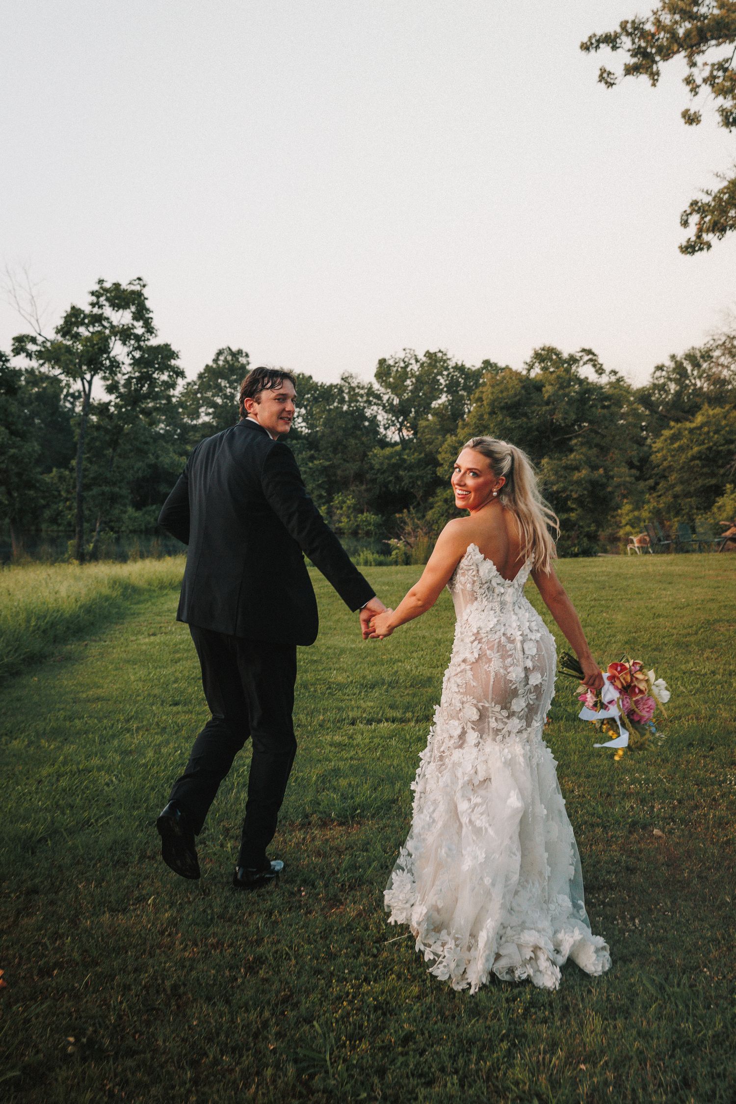 A couple in wedding attire walks playfully through a grassy field at sunset with trees in the background.
