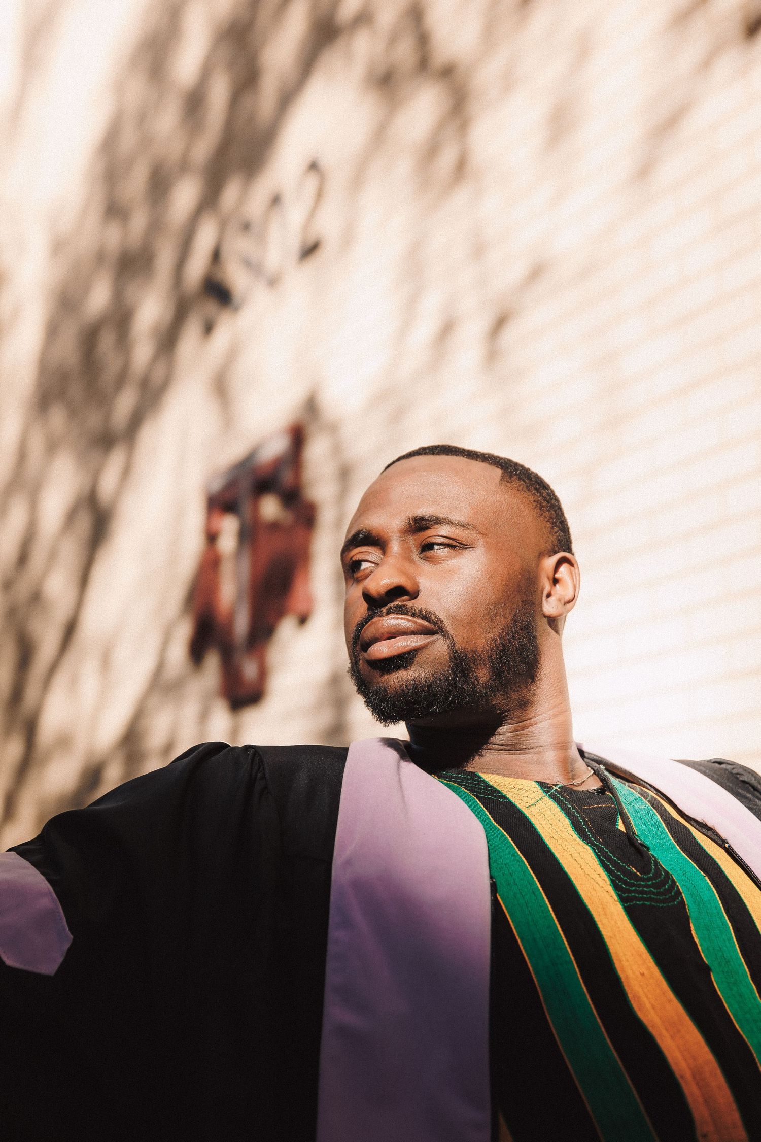 A college graduate with a beard wearing academic regalia featuring green and gold trim demonstrates different facial expressions.