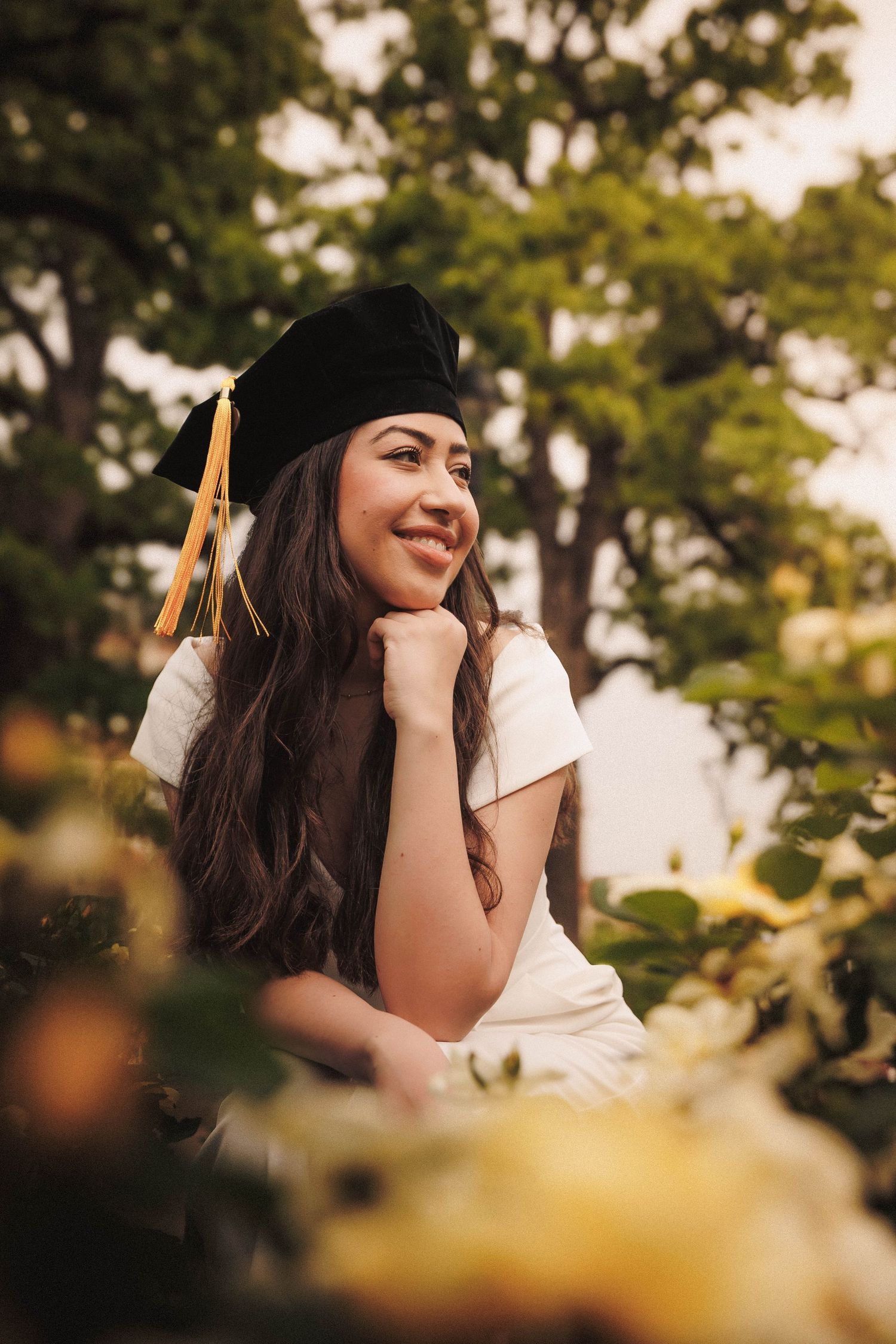 A graduate in a cap and gown smiles thoughtfully while posing outdoors among lush foliage during a summer photoshoot.