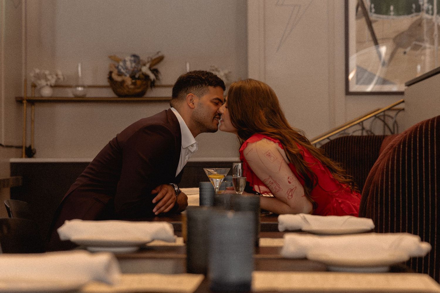 Couple sharing a romantic kiss at an elegant restaurant dinner table, man in suit and woman in red dress.