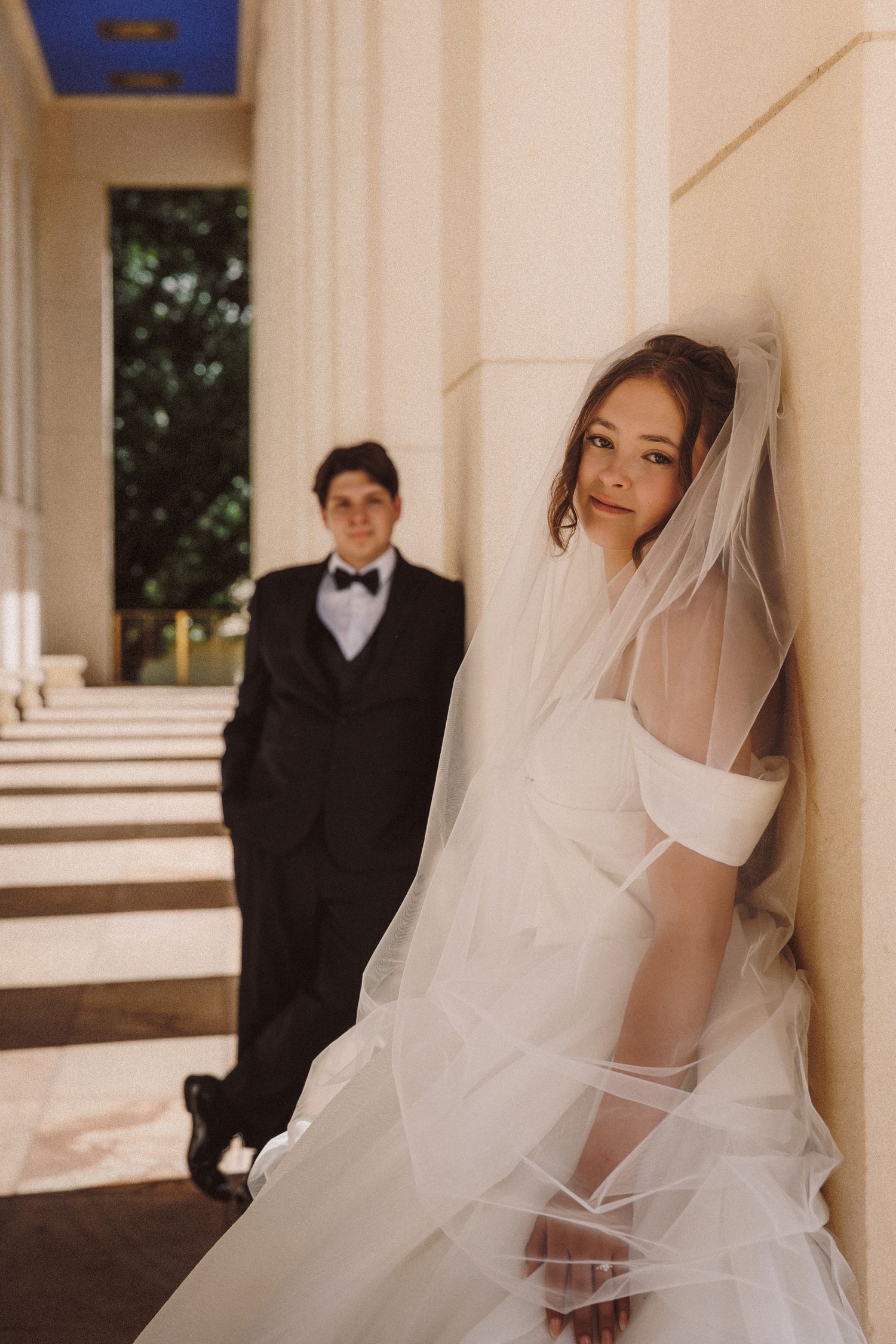 Bride in white veil leans against column while groom in tuxedo walks toward her.