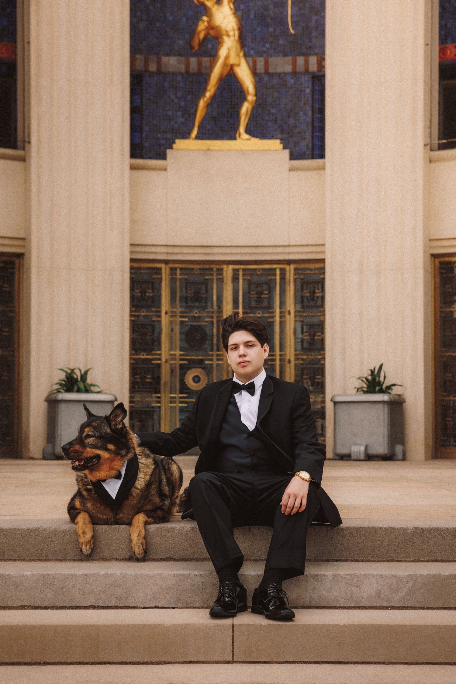 Groom in black tuxedo sits on steps with German Shepherd dog wearing bow tie at elegant venue with gold statue.