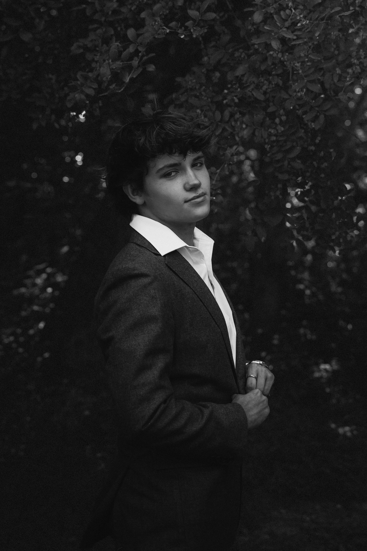 Artistic black and white photo of a figure in formal attire against a dark foliage background.