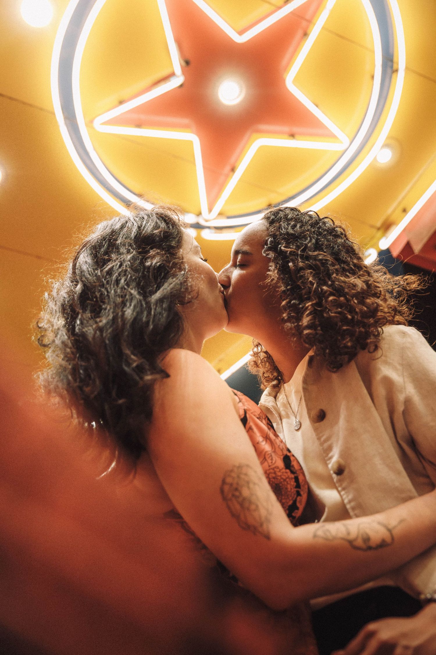 Two women share a romantic kiss beneath a glowing neon star sign inside a vintage theater venue.