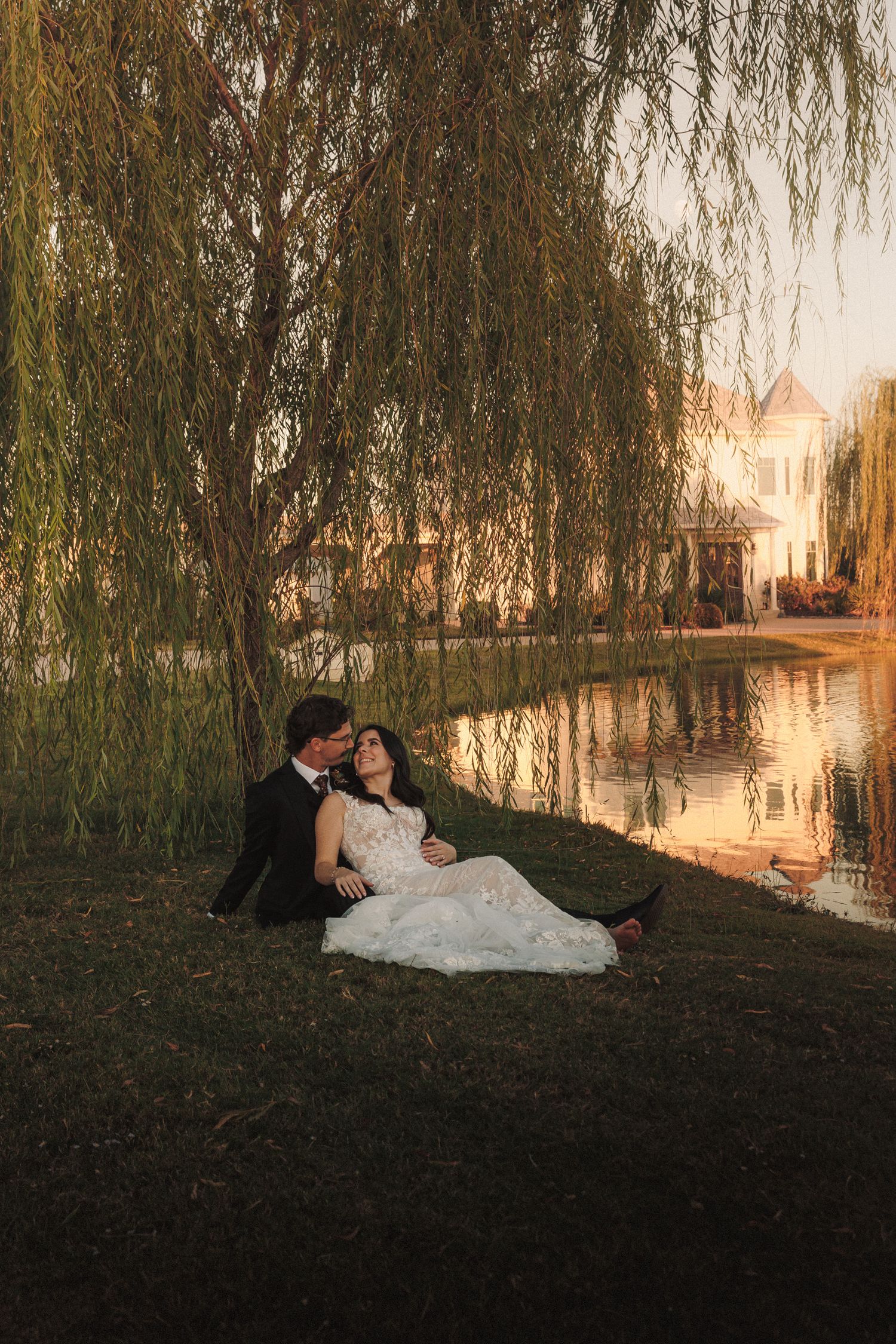Bride and groom sit romantically under a weeping willow tree by a reflective pond at sunset.