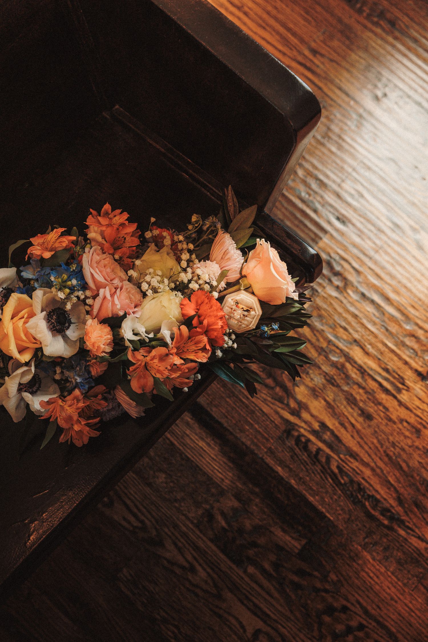 Colorful autumn wedding bouquet with orange roses, pink blooms, and baby's breath resting on a dark wooden church pew.