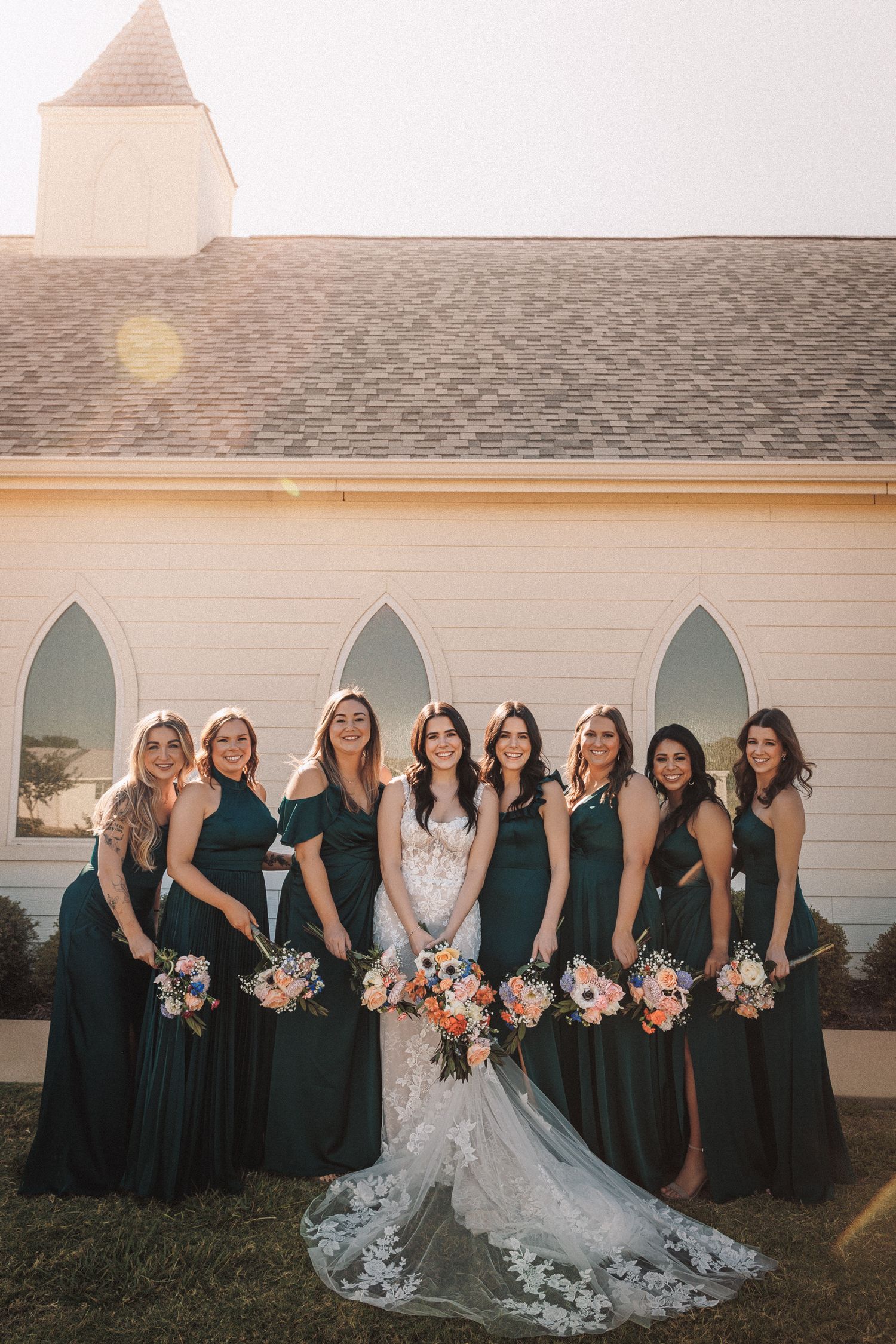 Bride in lace gown poses with seven bridesmaids in emerald green dresses holding bouquets outside white chapel.