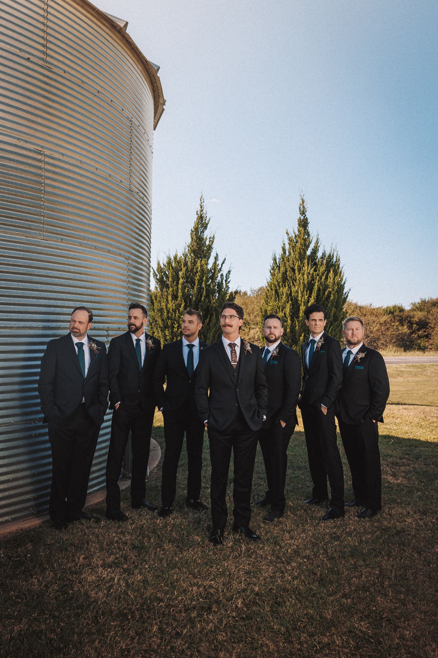 Groom and groomsmen in black suits pose together outdoors near a rustic grain silo on a sunny wedding day.