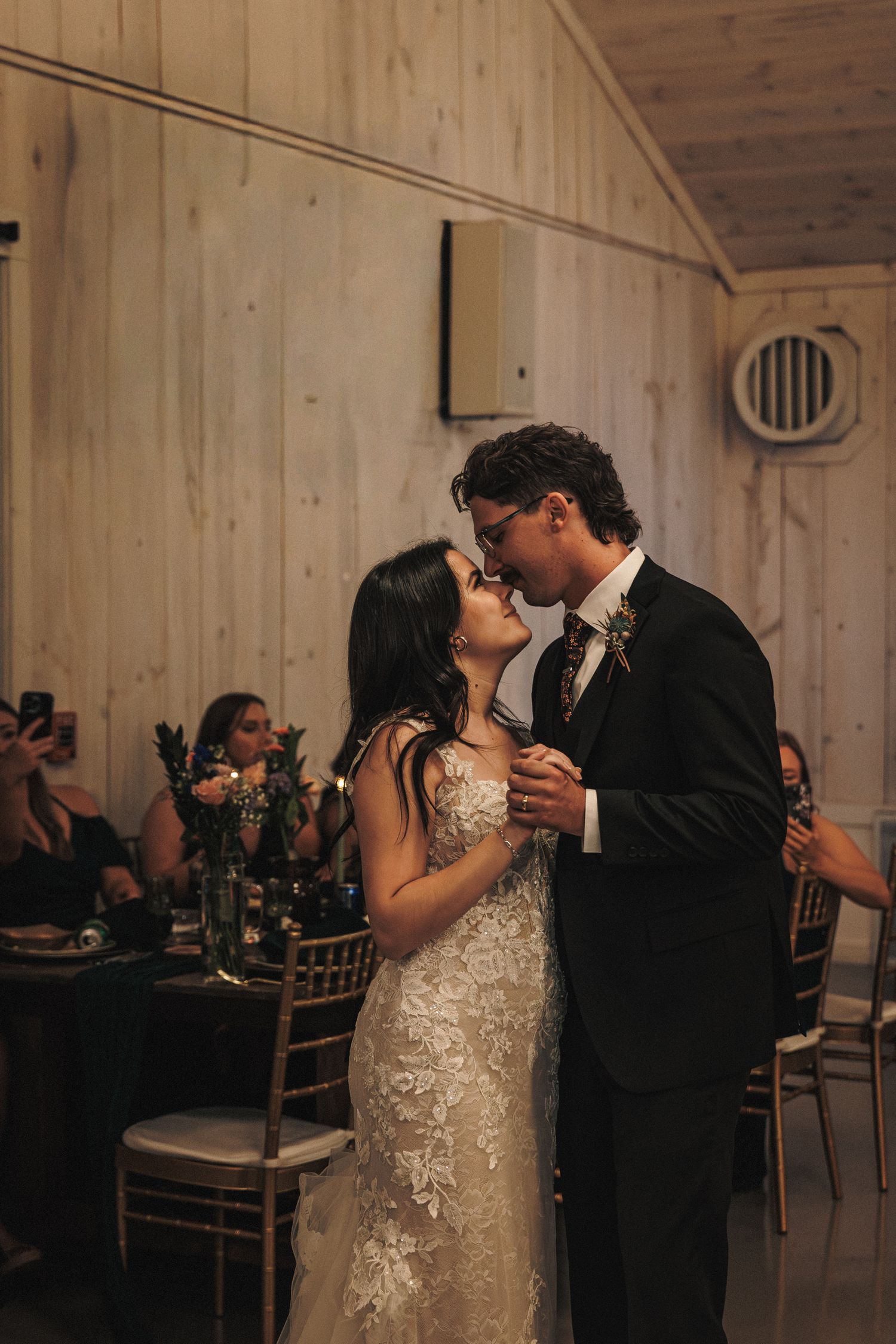 Bride and groom share their first dance kiss while guests celebrate in a warmly lit barn venue.