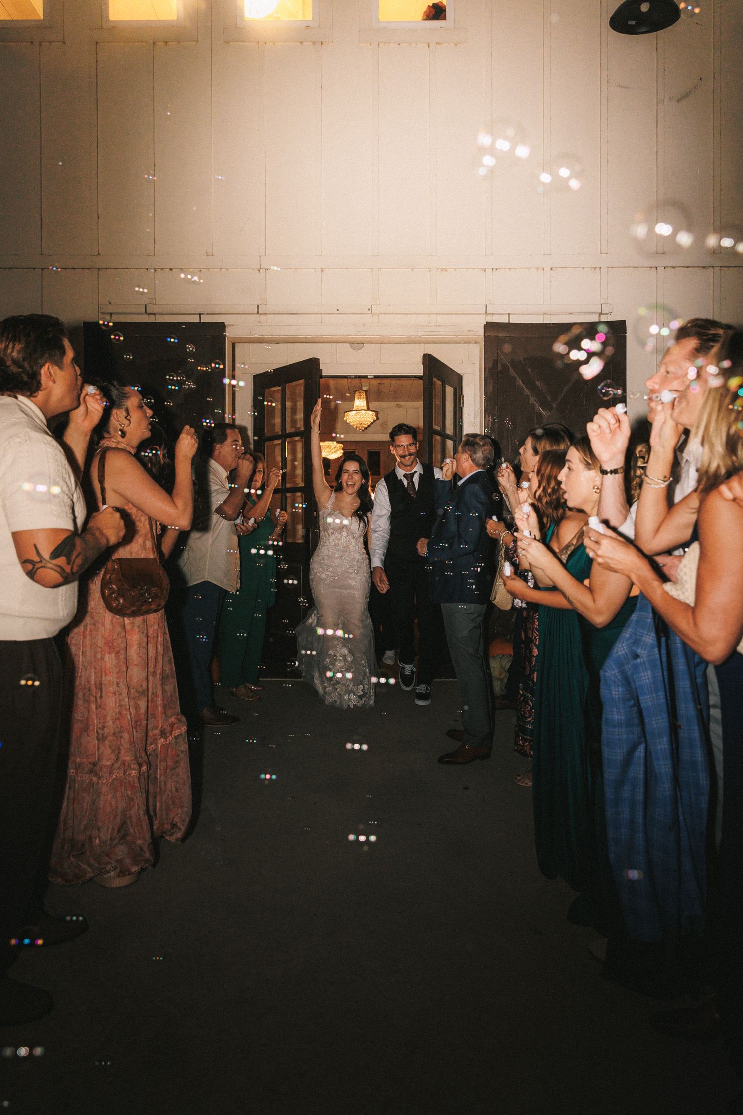 Bride and groom exit through bubble send-off surrounded by cheering guests at wedding reception.