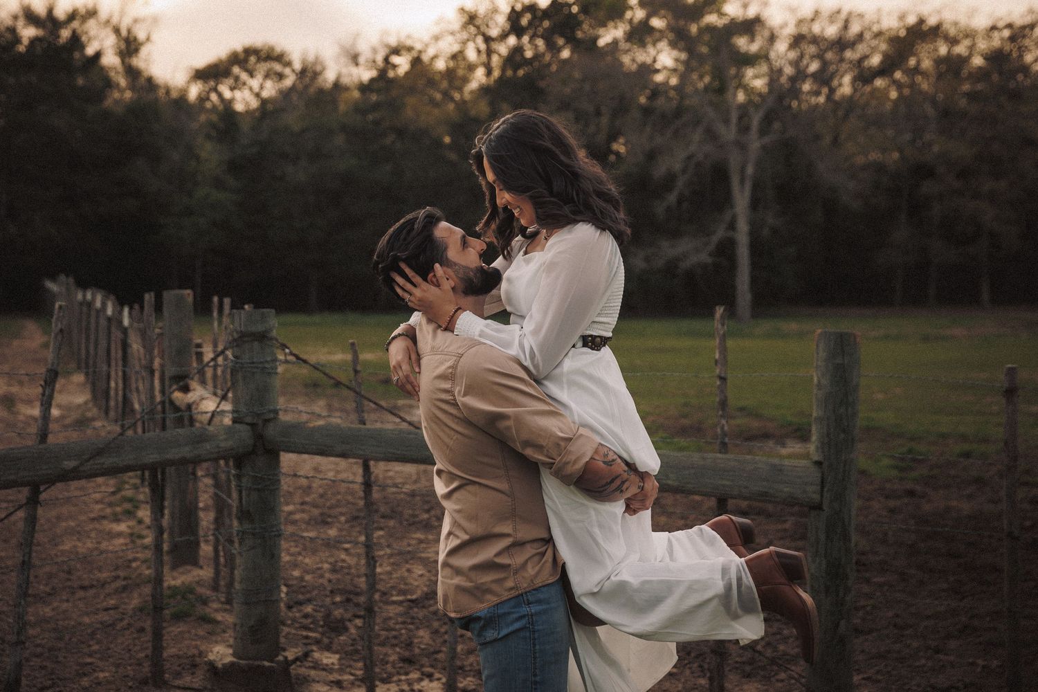 Man lifting woman in white dress at golden hour near a wooden farm fence.