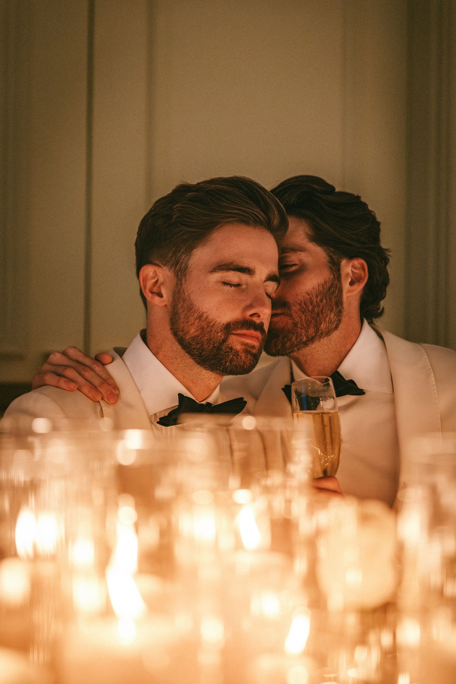 Two grooms share an intimate moment surrounded by glowing candles and champagne glasses.