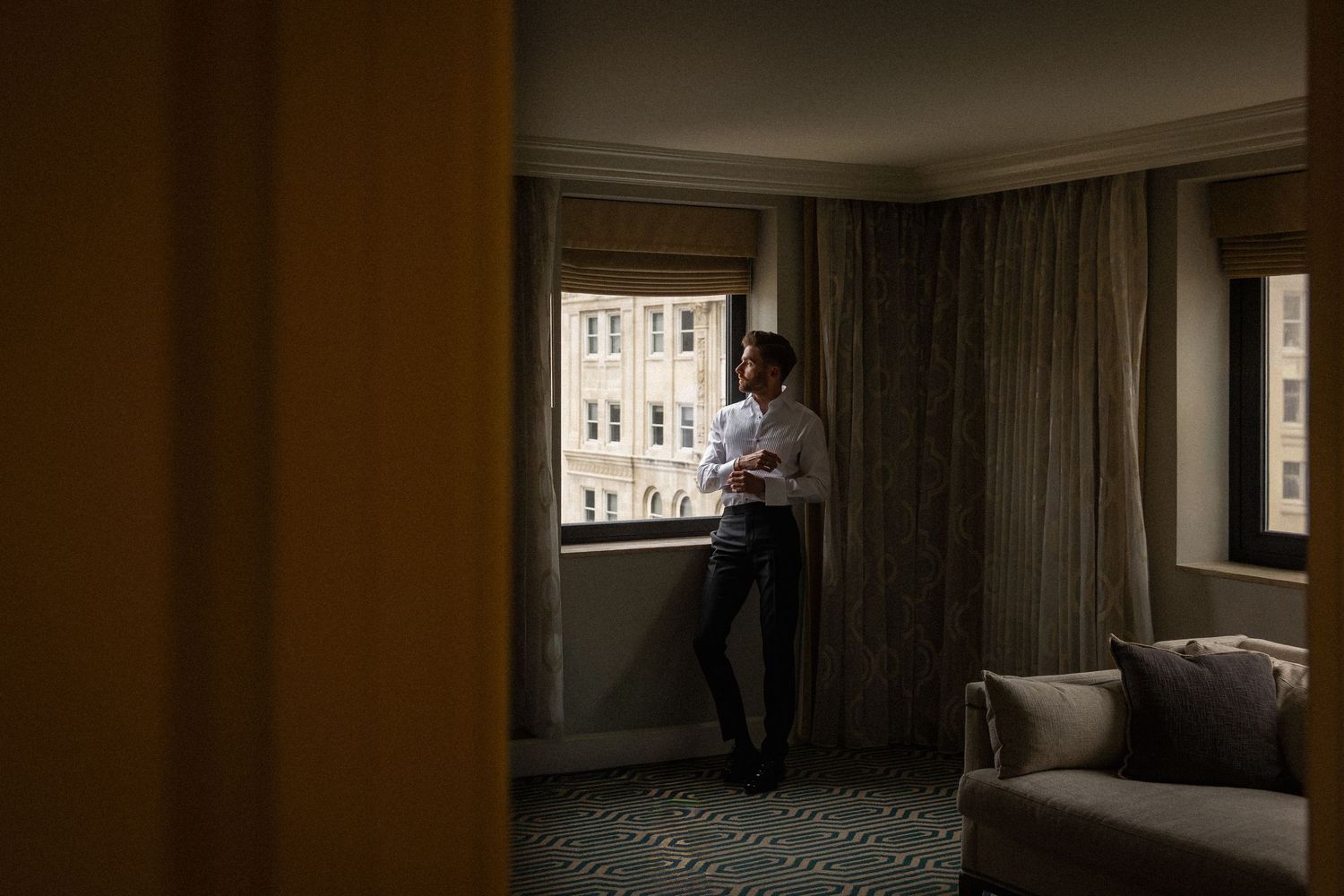 A man in a white shirt stands contemplatively by a hotel room window with city views.