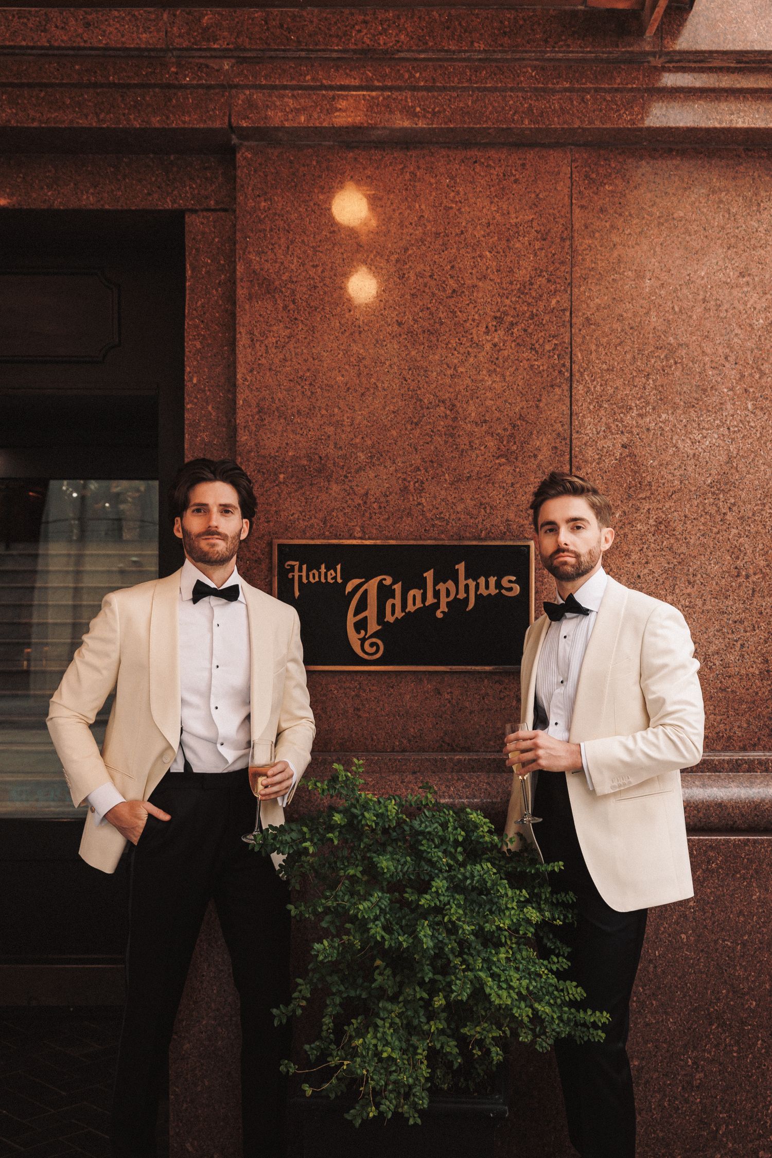 Two grooms in ivory tuxedos with bow ties pose outside Hotel Adolphus for their wedding day.
