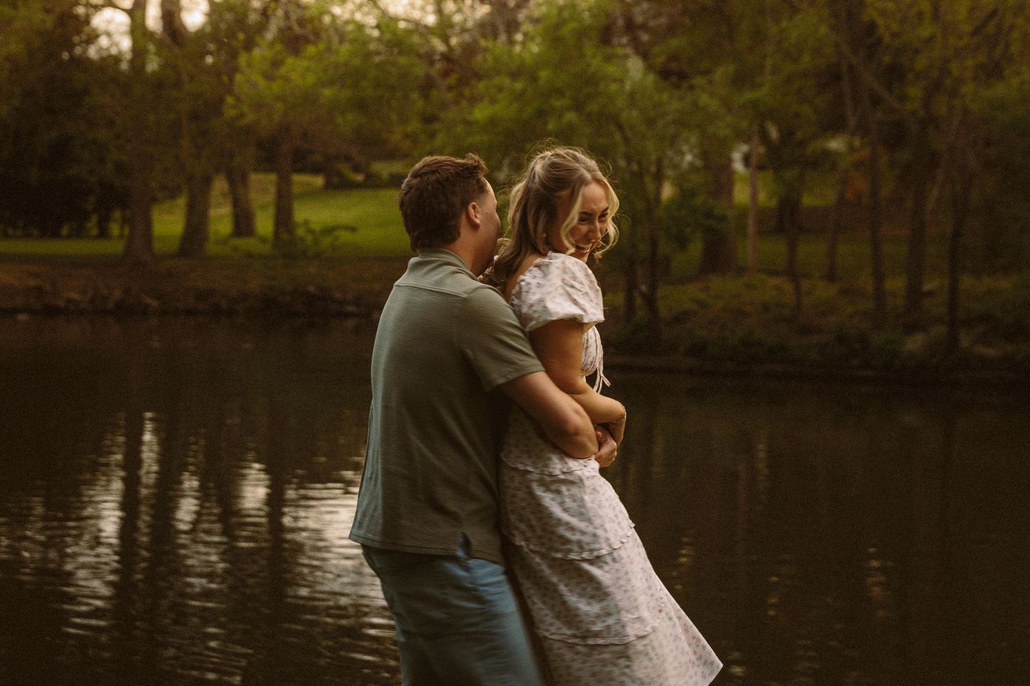 Couple embracing by a reflective pond at dusk, surrounded by lush green trees.