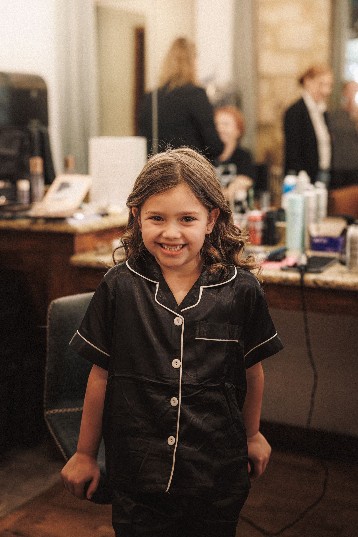 Adorable young girl in black pajamas smiling in bridal suite during wedding hair and makeup preparations.