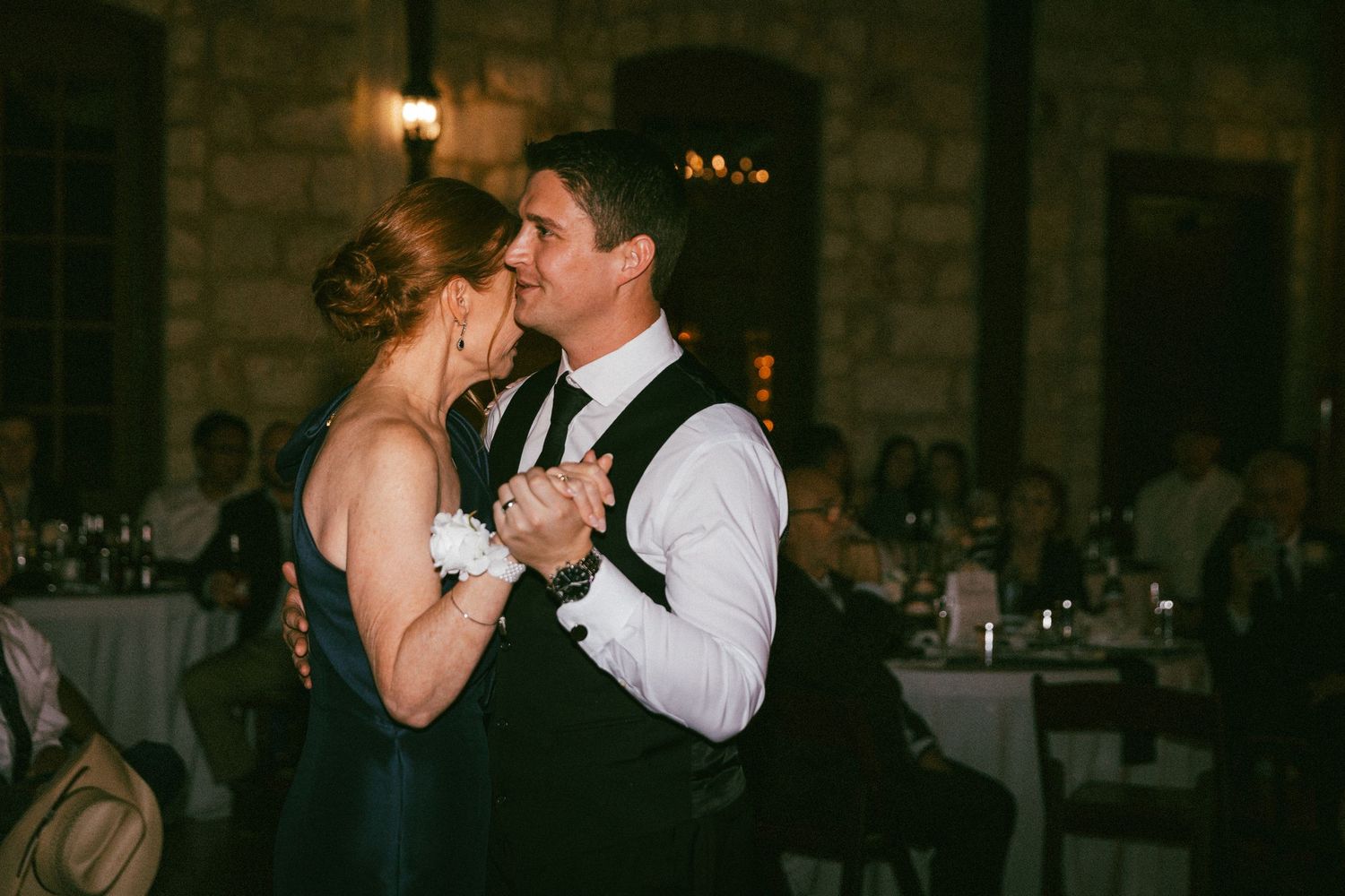 Groom and mother share emotional first dance in dimly lit rustic reception venue.