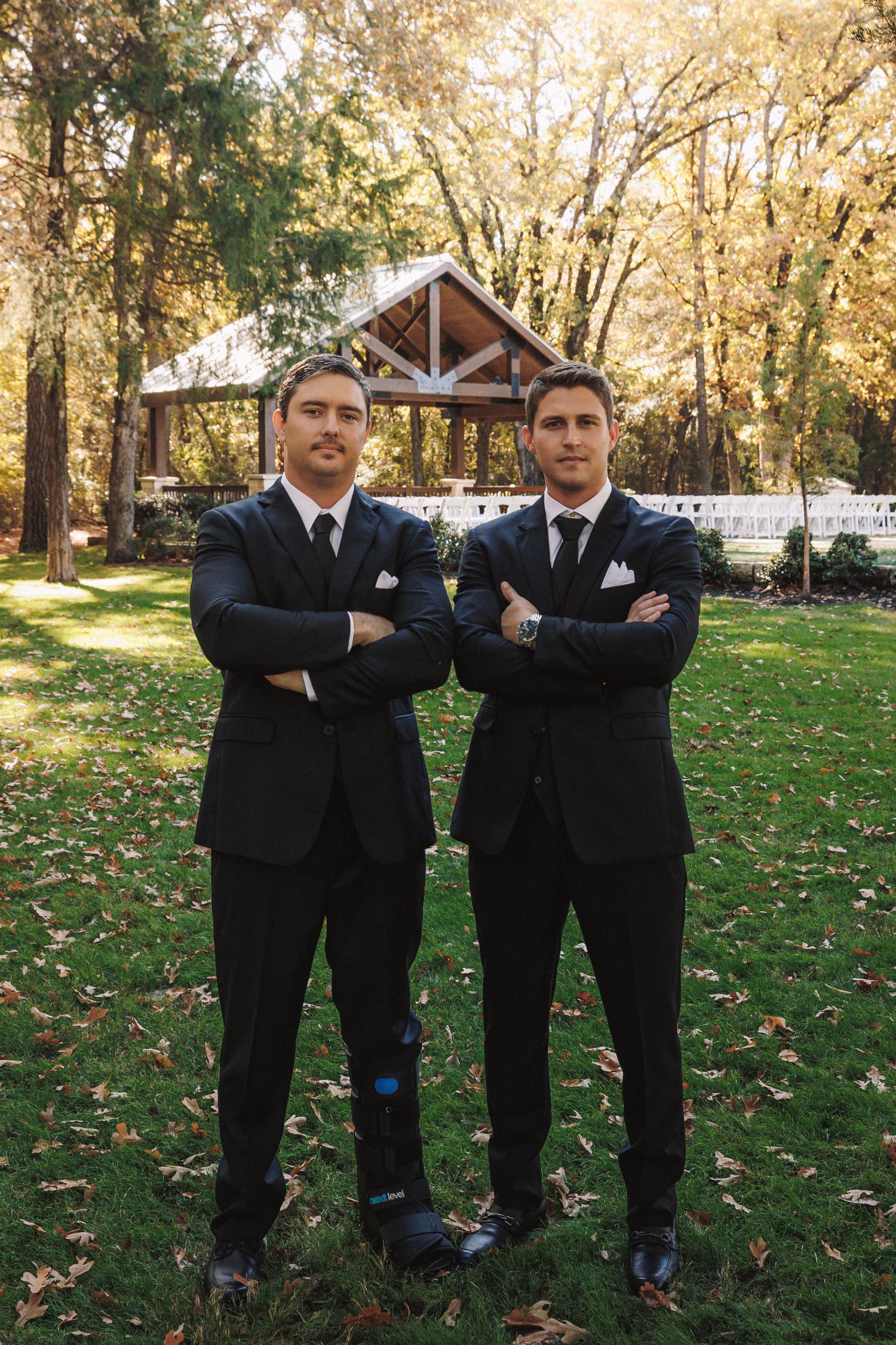 Two groomsmen in black suits with arms crossed standing before an outdoor wedding ceremony pavilion.