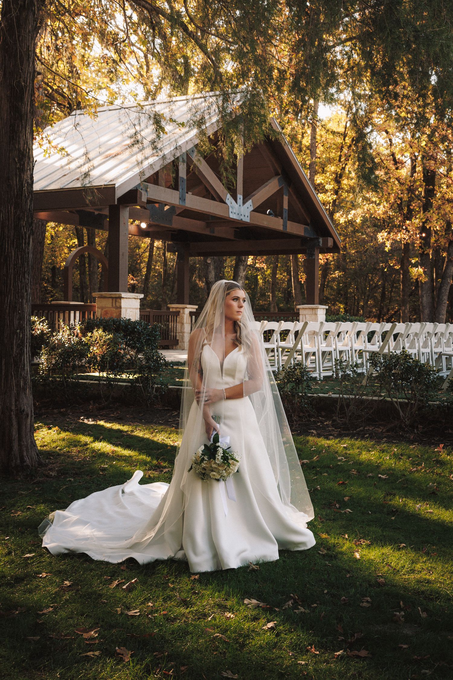 Elegant bride in white gown and veil standing near a wooden gazebo in an autumn forest setting.