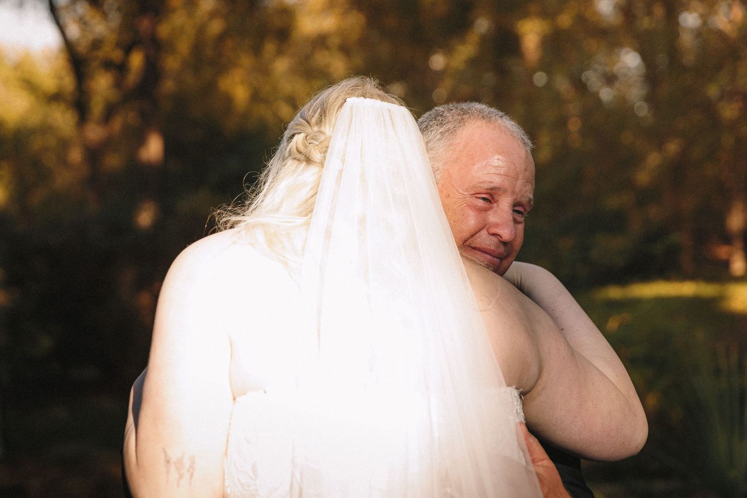Emotional father hugging his daughter in a wedding dress and veil in an outdoor autumn setting.