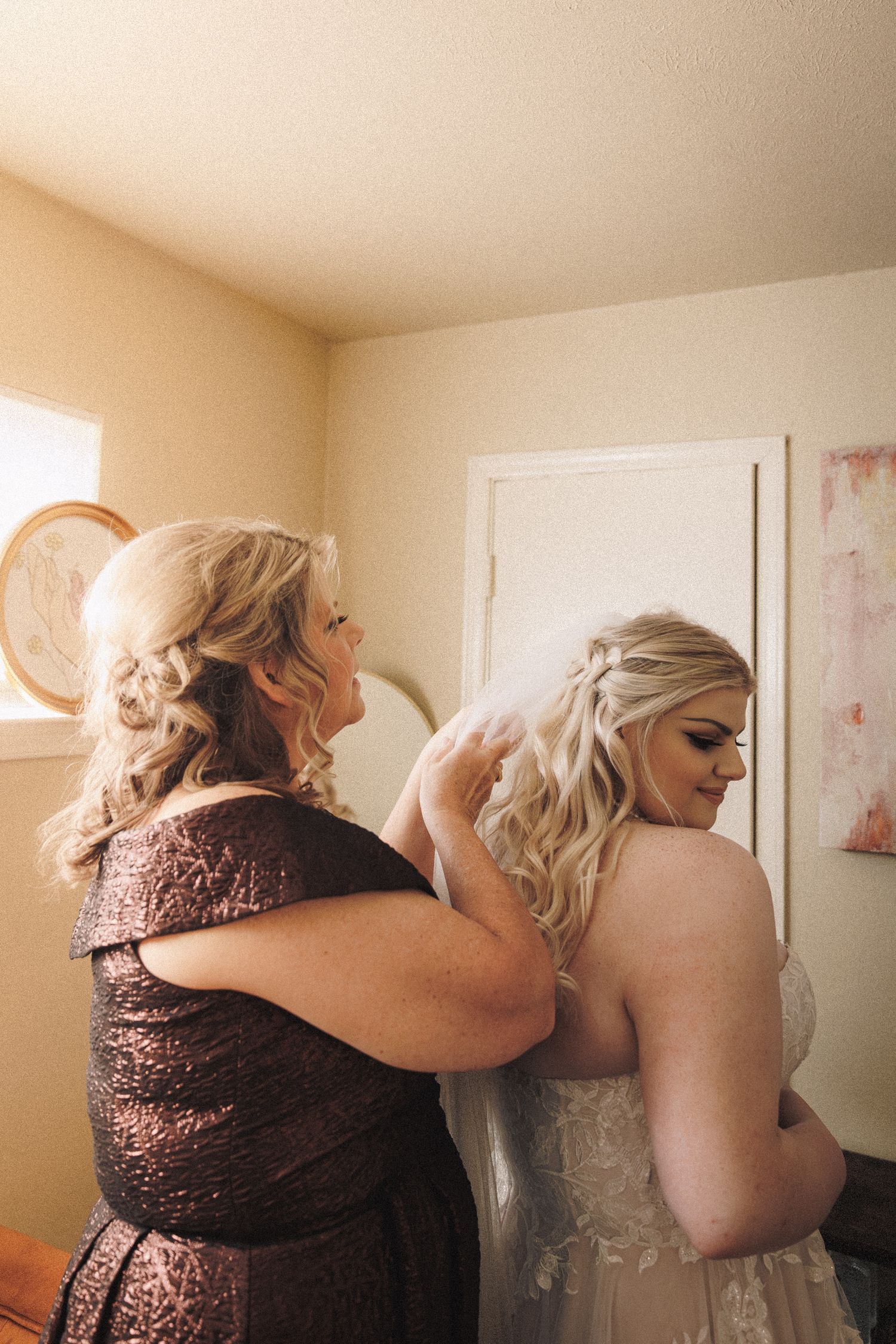 Mother helping bride with veil during wedding morning preparations indoors.