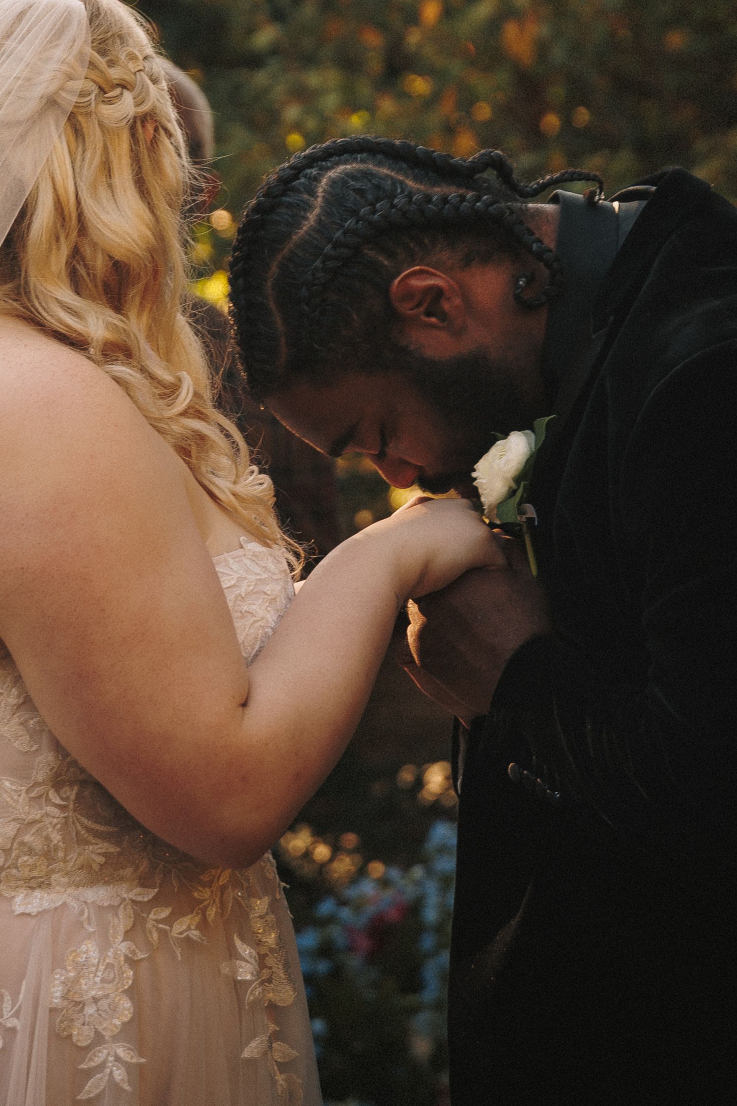 Groom in black tuxedo kissing bride's hand during outdoor wedding ceremony at golden hour.