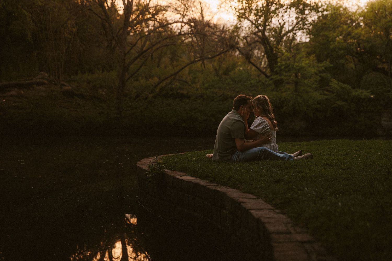 Couple sitting close together by a pond at sunset in a lush green park, sharing a romantic moment.