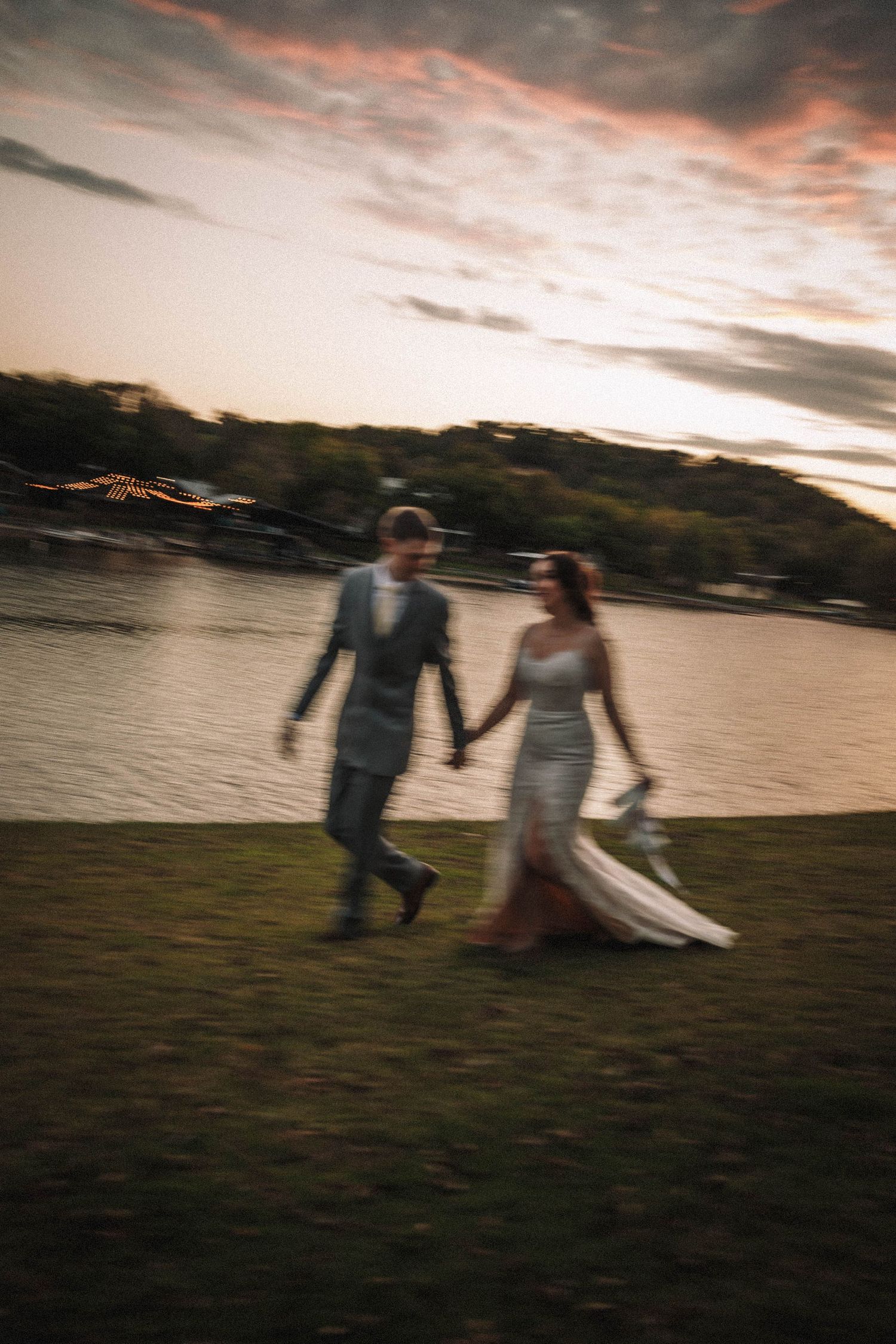 A couple in wedding attire walks hand in hand along a lakeside at sunset with dramatic evening clouds overhead.