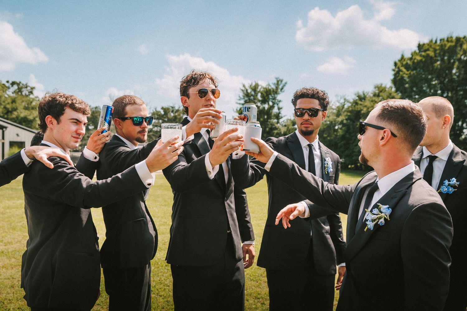 Groomsmen in black suits toasting drinks outdoors during wedding celebration.