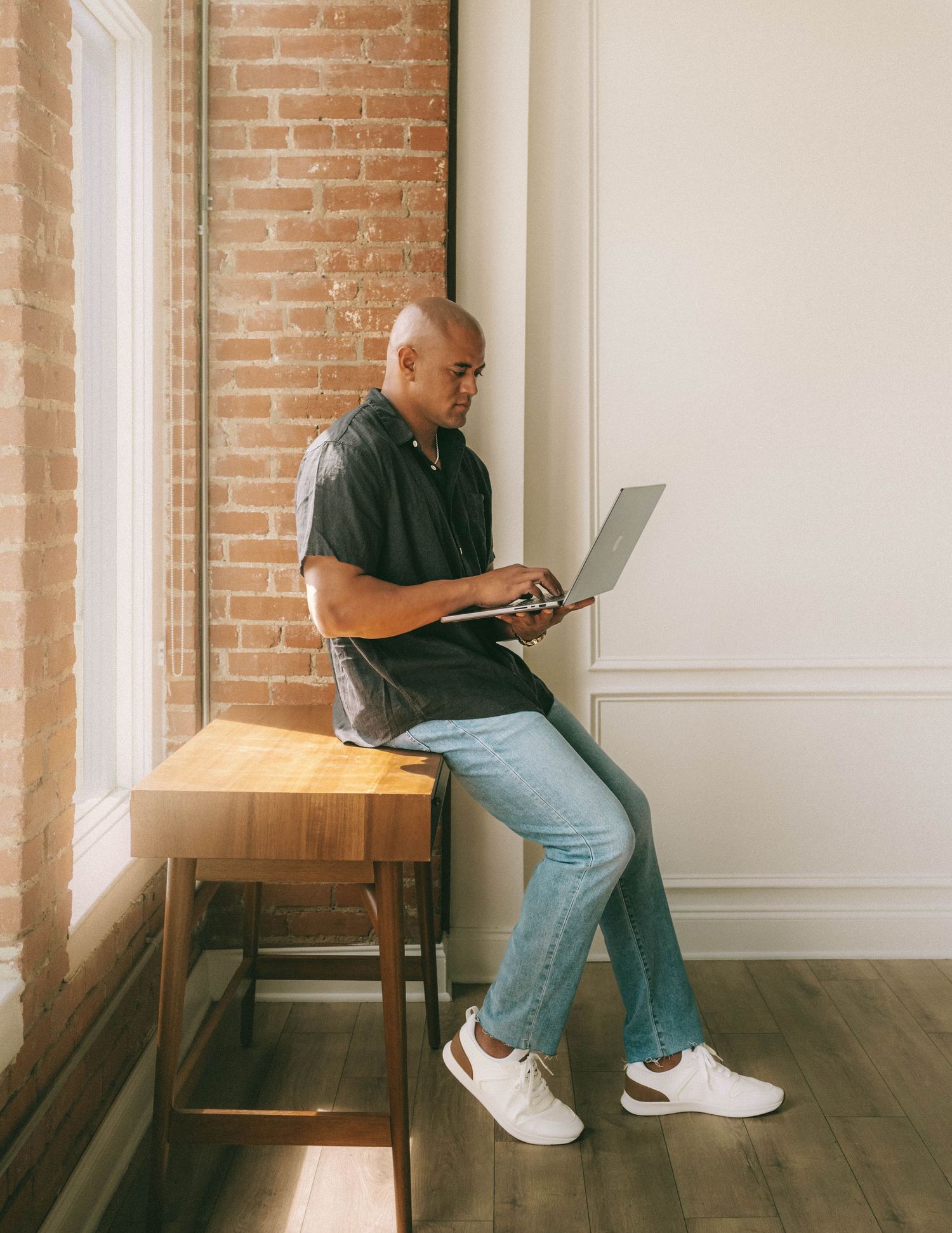 Someone in jeans and a dark shirt sits with laptop on wooden stool against exposed brick wall in modern space.