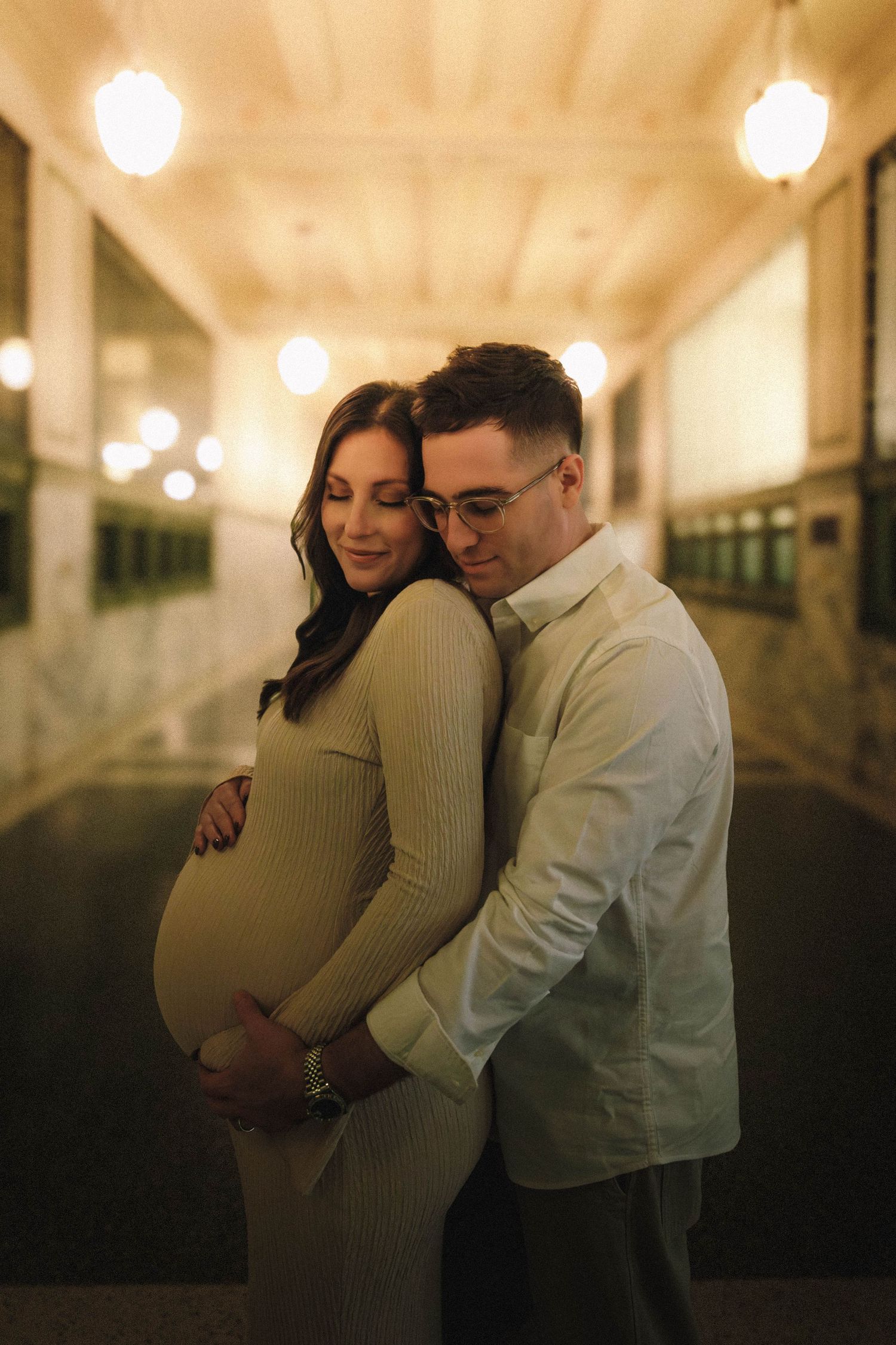 Couple embraces tenderly in a romantic maternity photoshoot with soft glowing lights in the background.