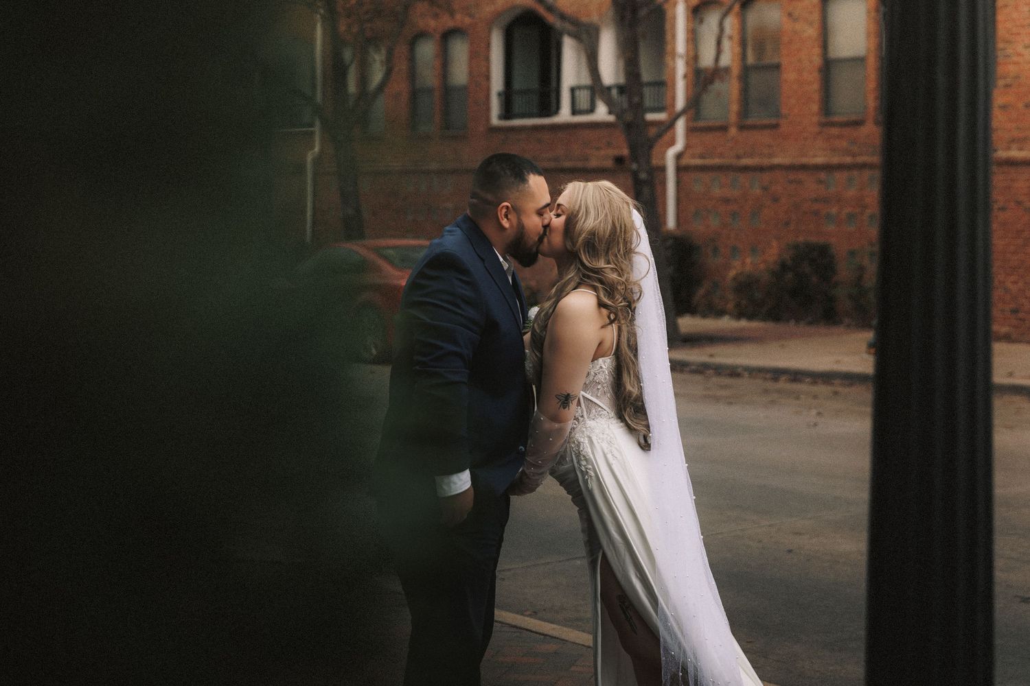 Newlywed couple embraces in romantic kiss on brick building background during outdoor wedding photoshoot.
