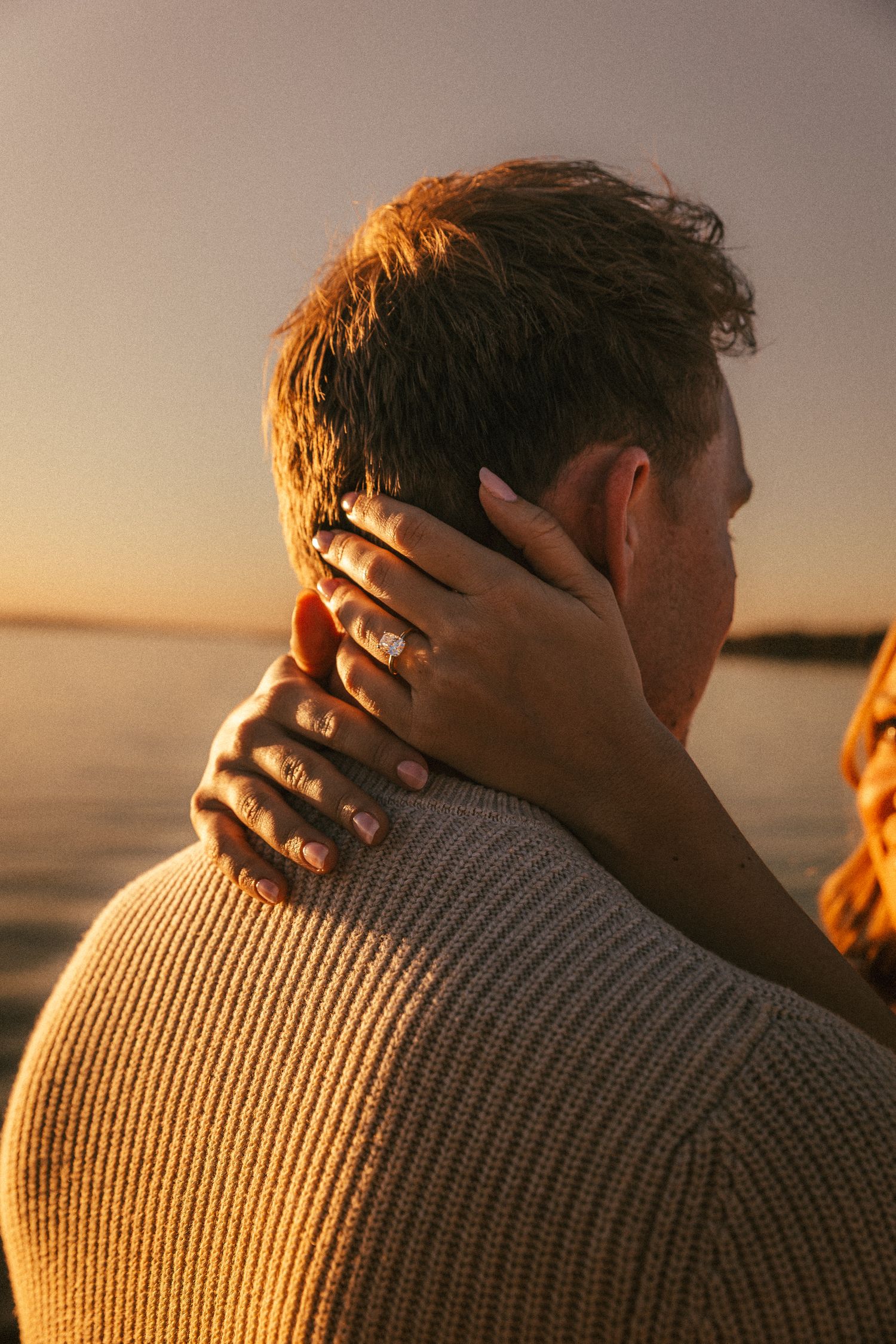 Intimate sunset moment with hands embracing neck and ring visible against warm golden light.