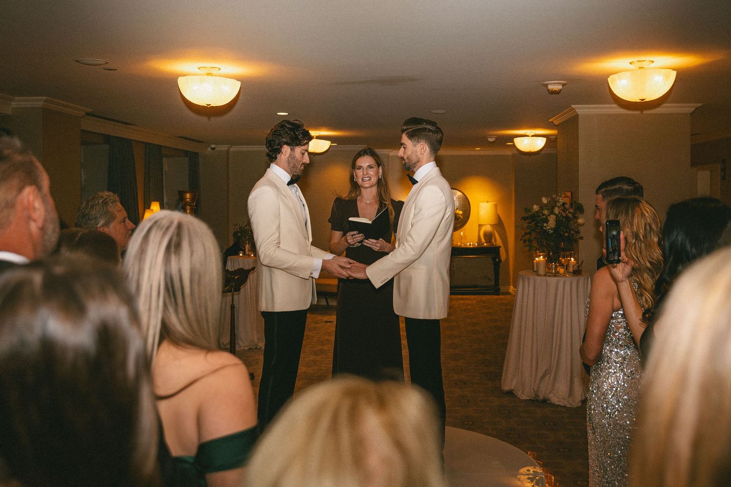 Two grooms in white tuxedos exchange vows with officiant during intimate wedding ceremony.