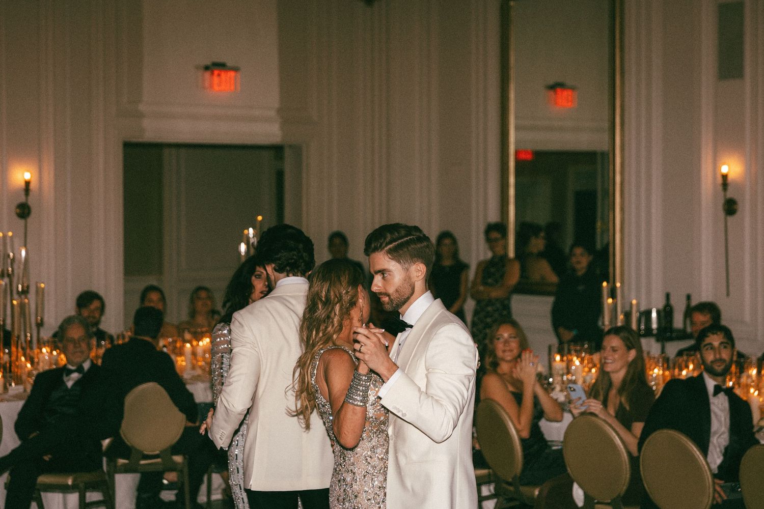 Groom in white tuxedo sharing a dance with a guest at a candlelit wedding reception.