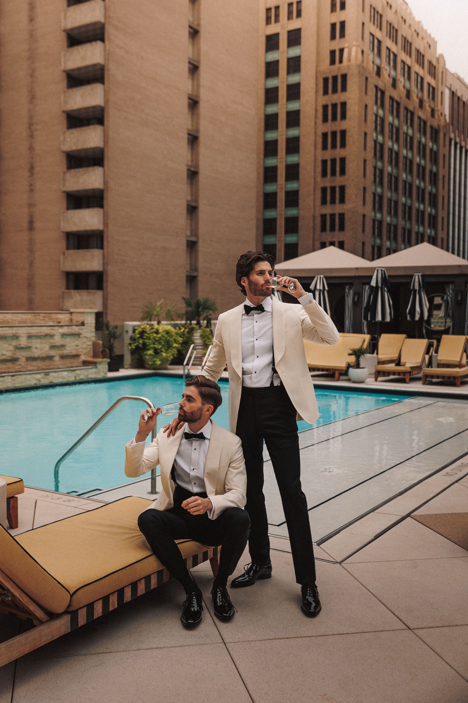 Two grooms in white tuxedo jackets enjoy drinks poolside at a luxury hotel rooftop.