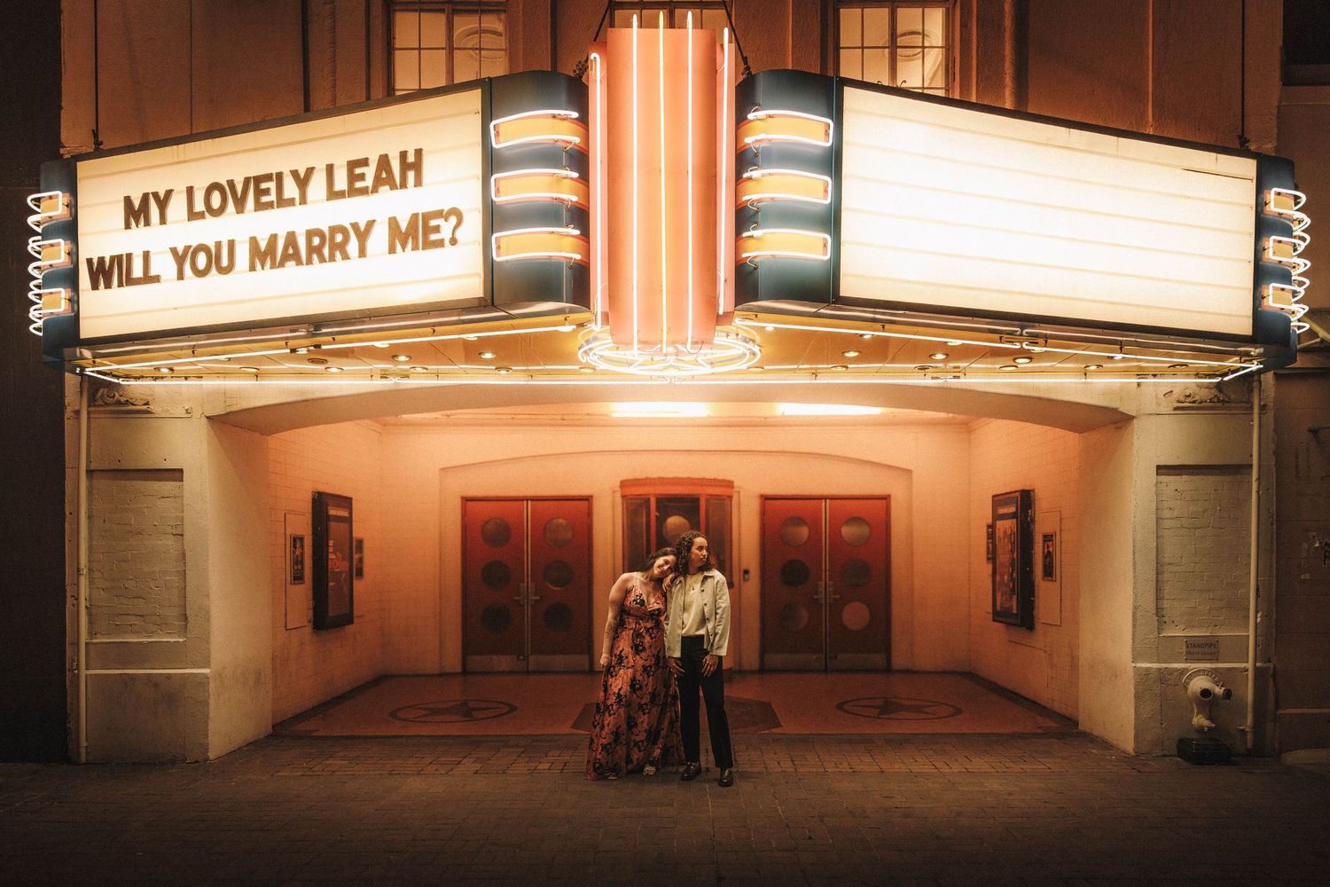 Newly engaged couple standing together under illuminated theater marquee with marriage proposal message at night.