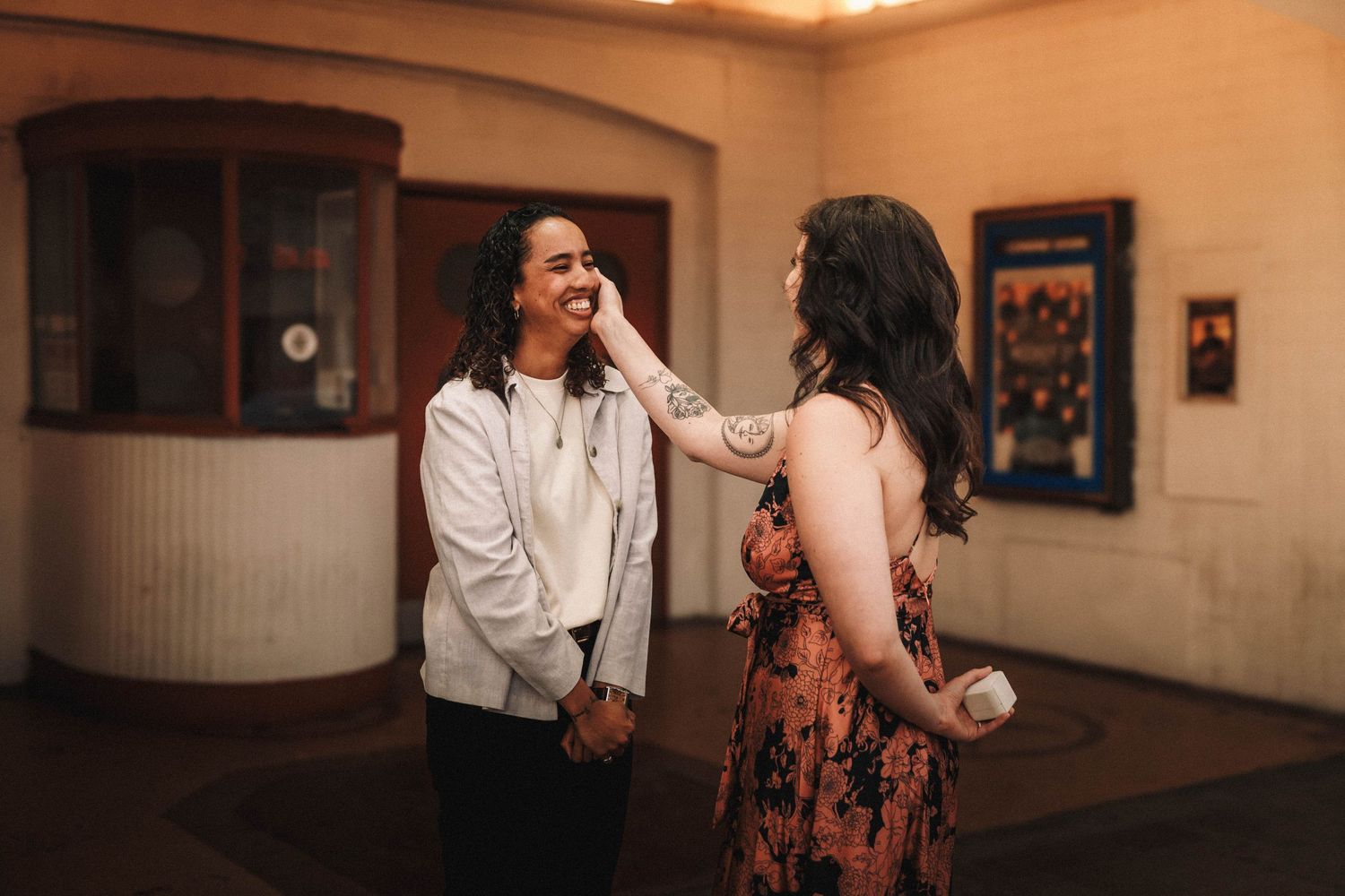Two women laughing and sharing a joyful moment in a warmly lit venue lobby, one touching the other's face.