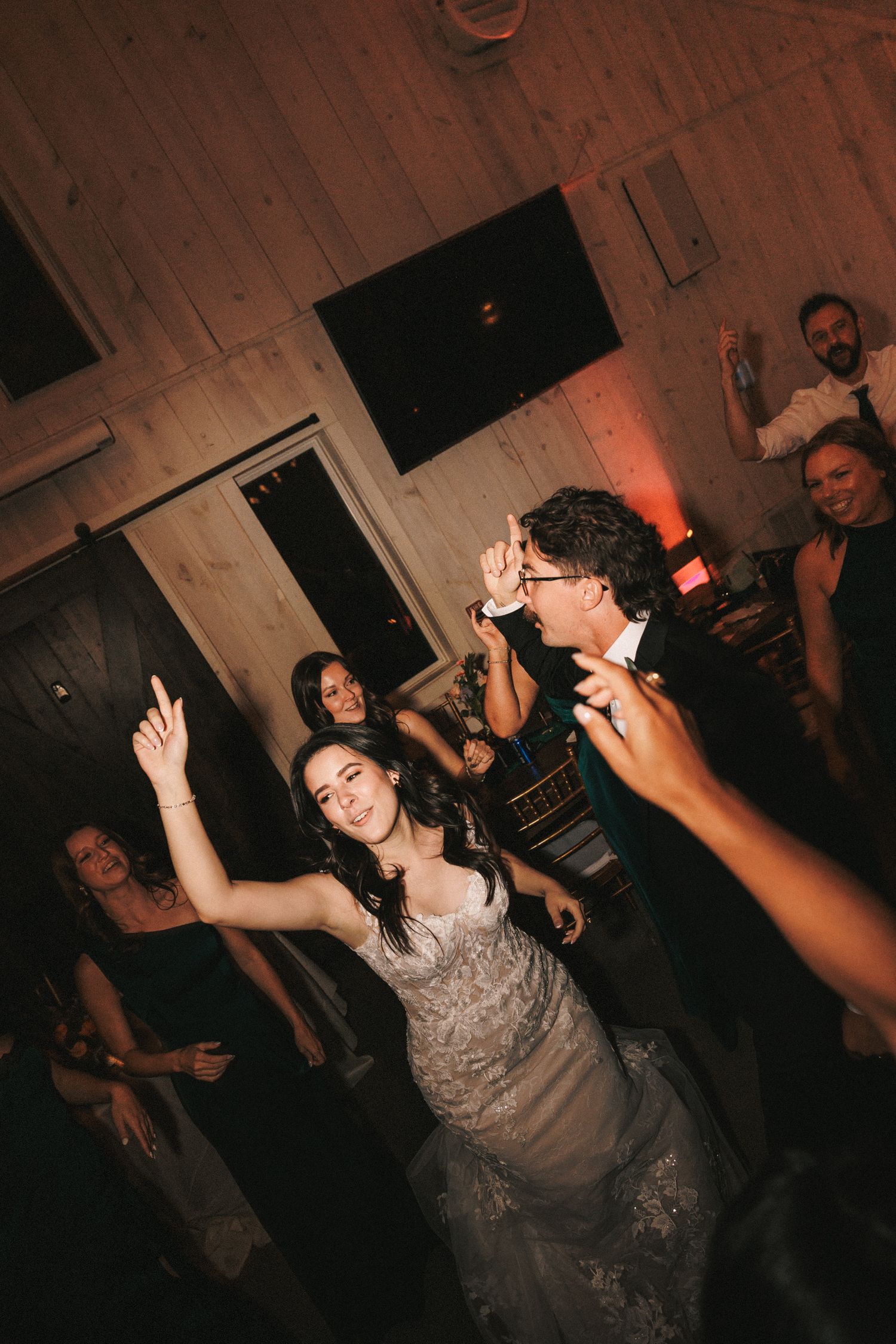 Bride in lace gown dancing joyfully with groom and guests on a warmly lit wedding reception dance floor.