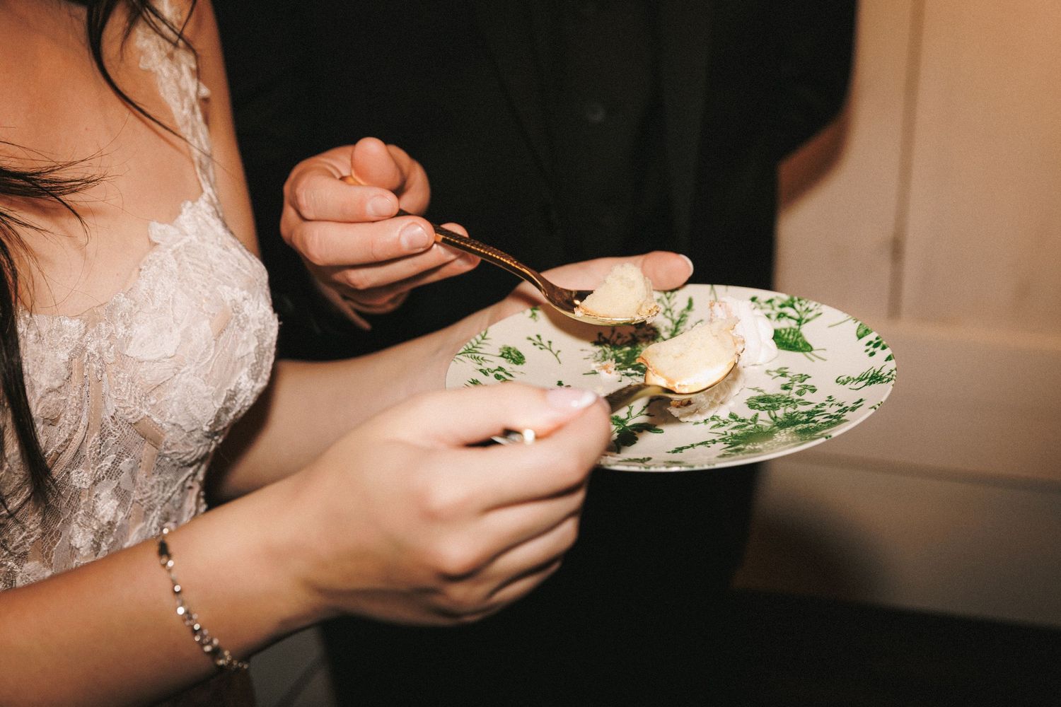 Bride holding green floral plate while groom serves wedding cake with gold spoon at reception.